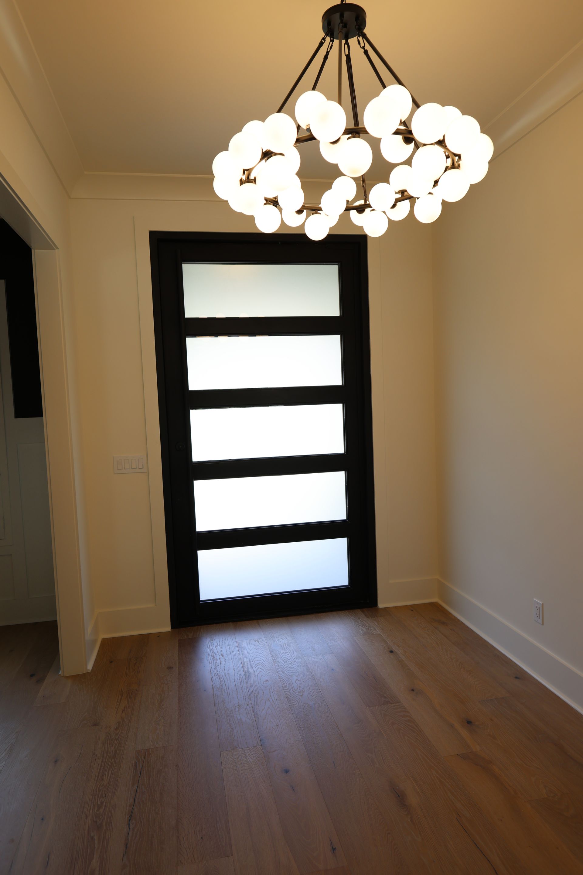 Black-framed door with frosted glass panels in an empty room, wooden floor, chandelier, and white walls.