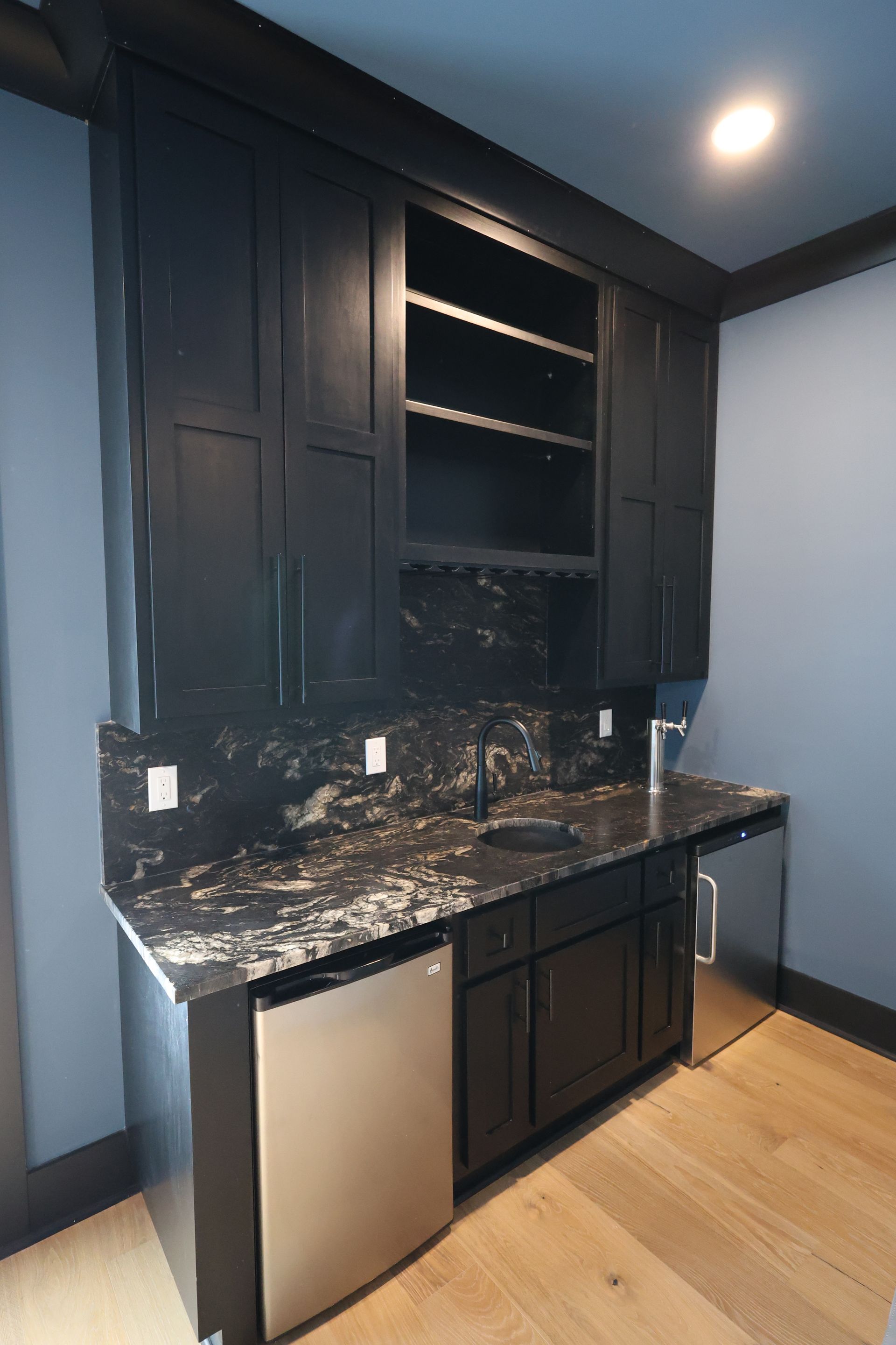 Black wet bar with marble countertop, cabinets, and appliances against a blue wall.