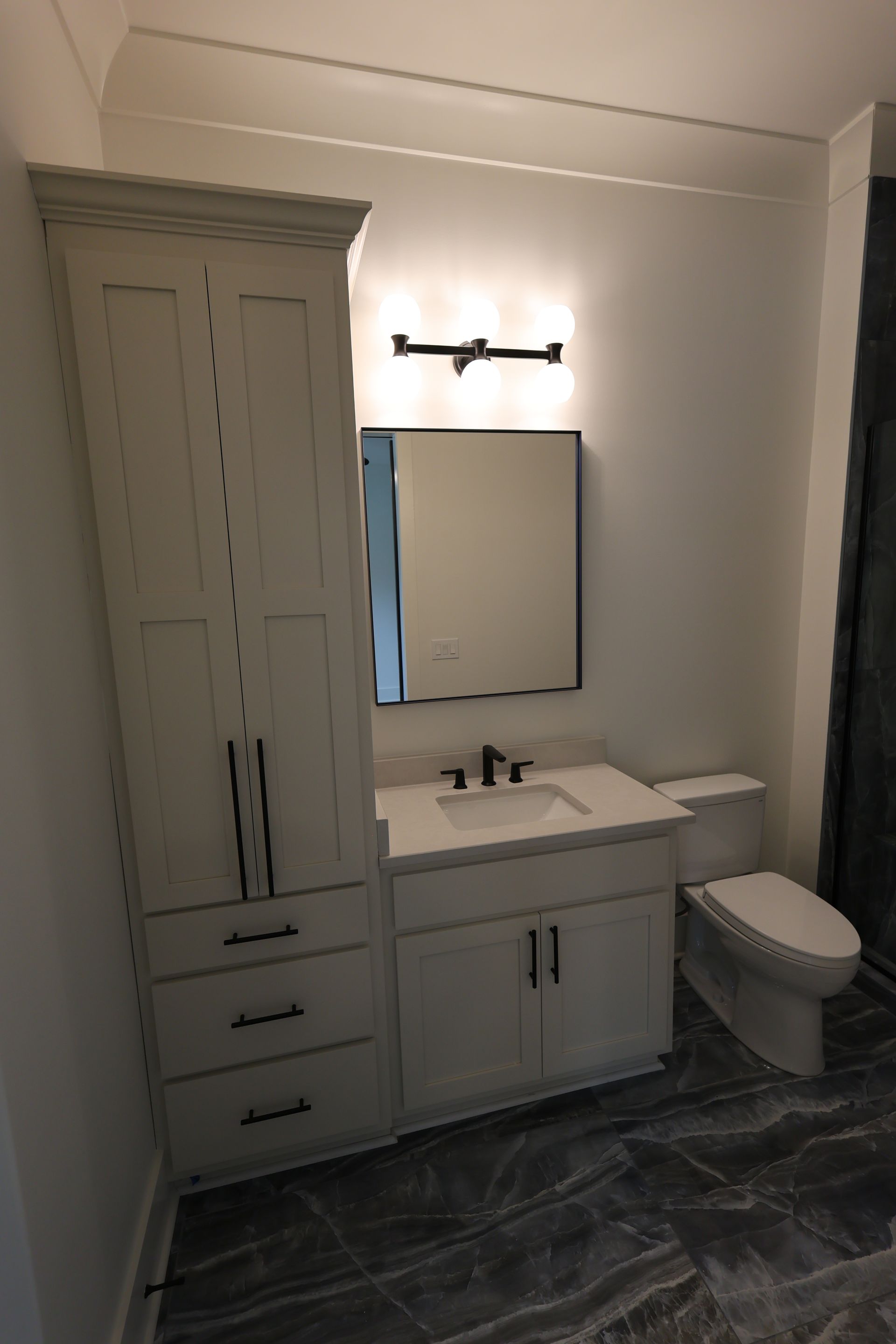 Bathroom with white vanity, tall storage cabinet, toilet, and black fixtures.