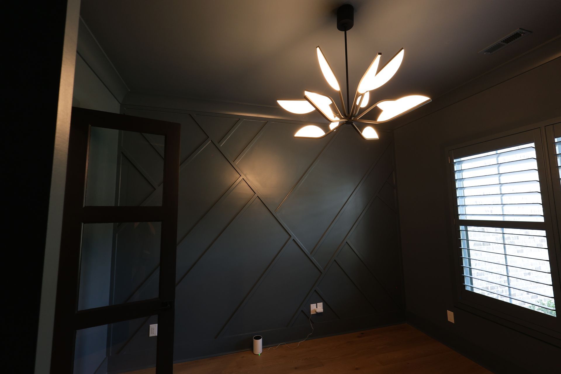 Dark-walled room with geometric paneling, modern chandelier, window with shutters, and a closed glass door.