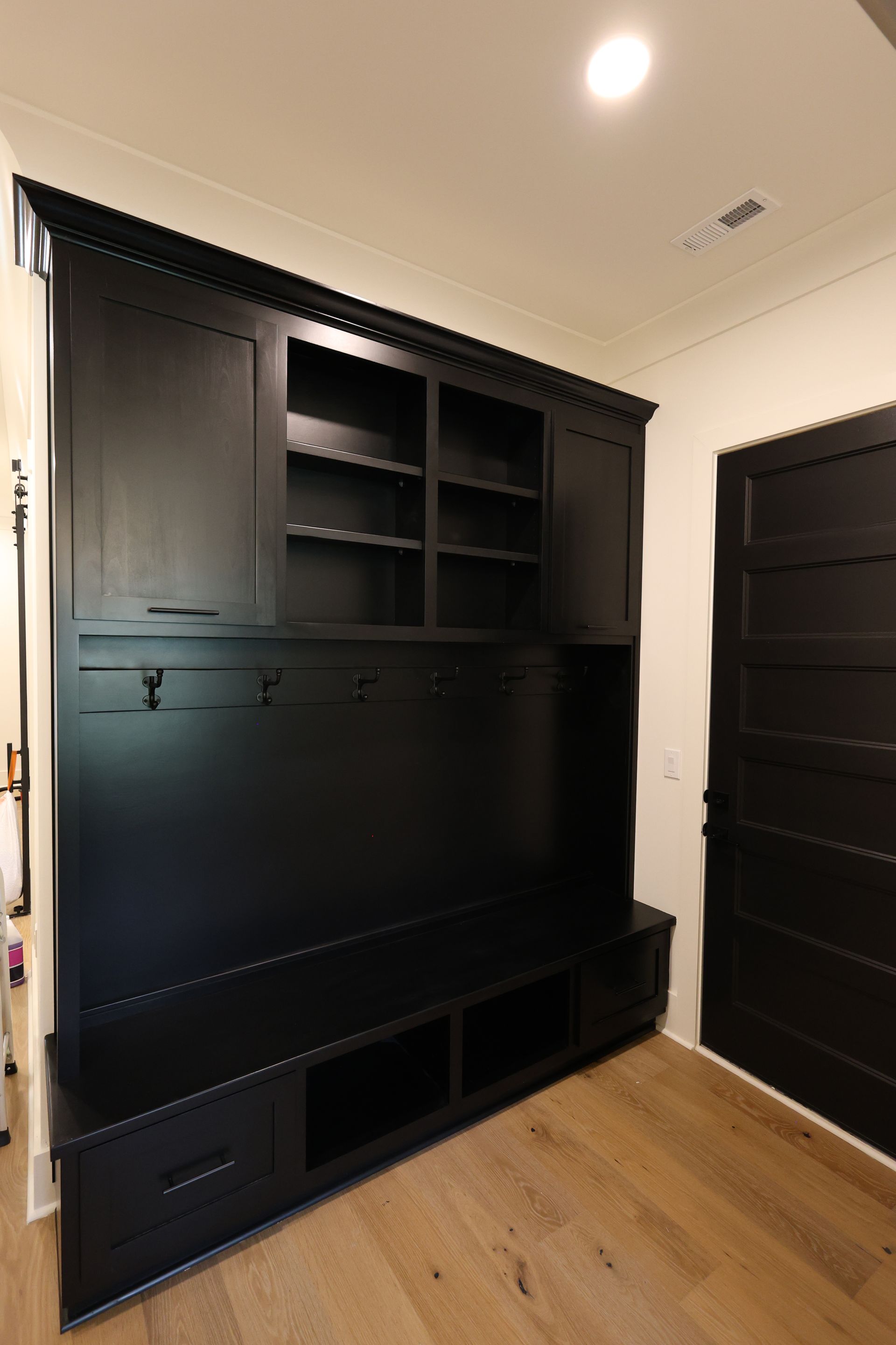 Black built-in entryway storage with shelves, bench, drawers, and hooks. Wooden floor. Door on the right.
