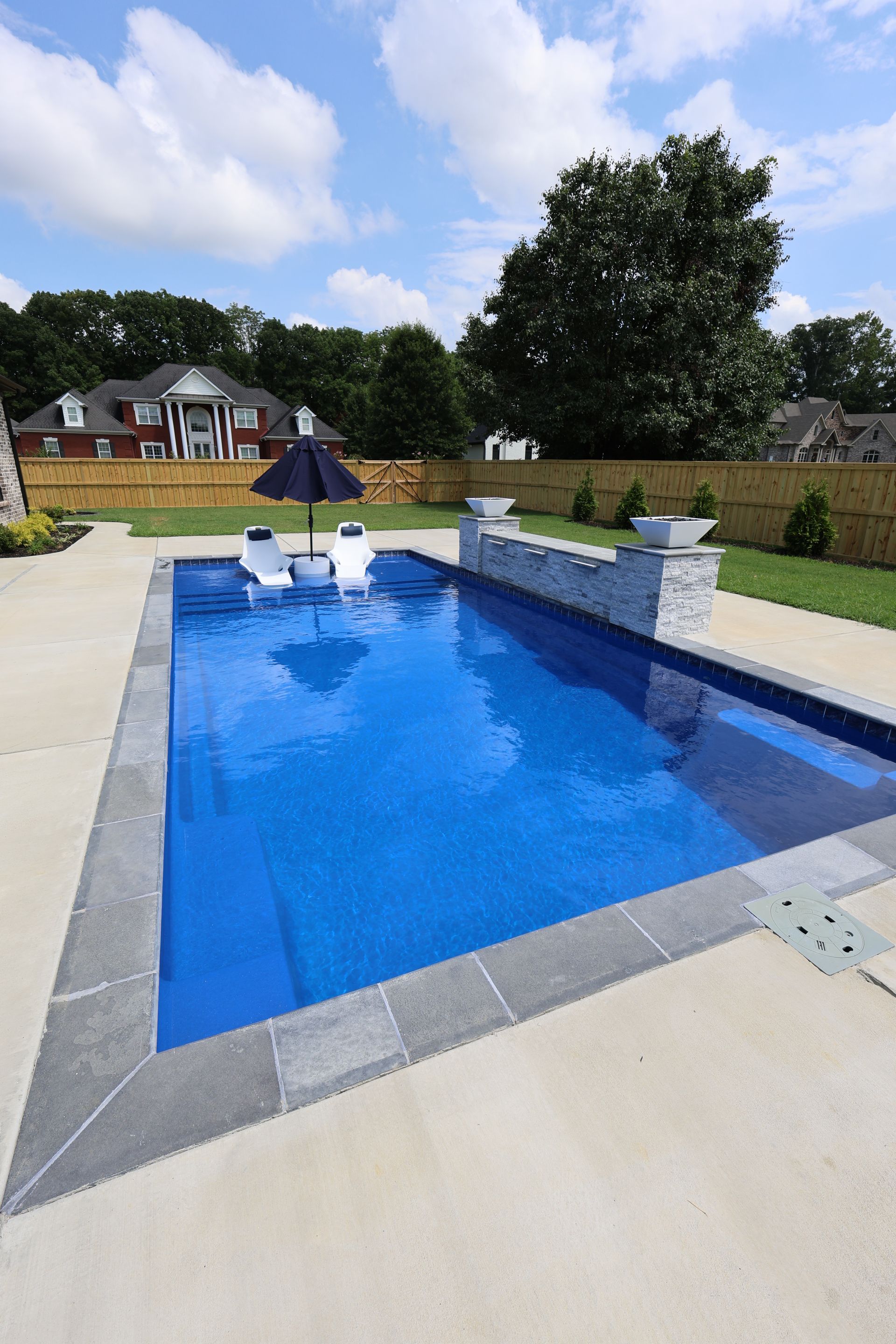 Rectangular blue swimming pool in backyard with lounge chairs, blue umbrella, and stone patio.