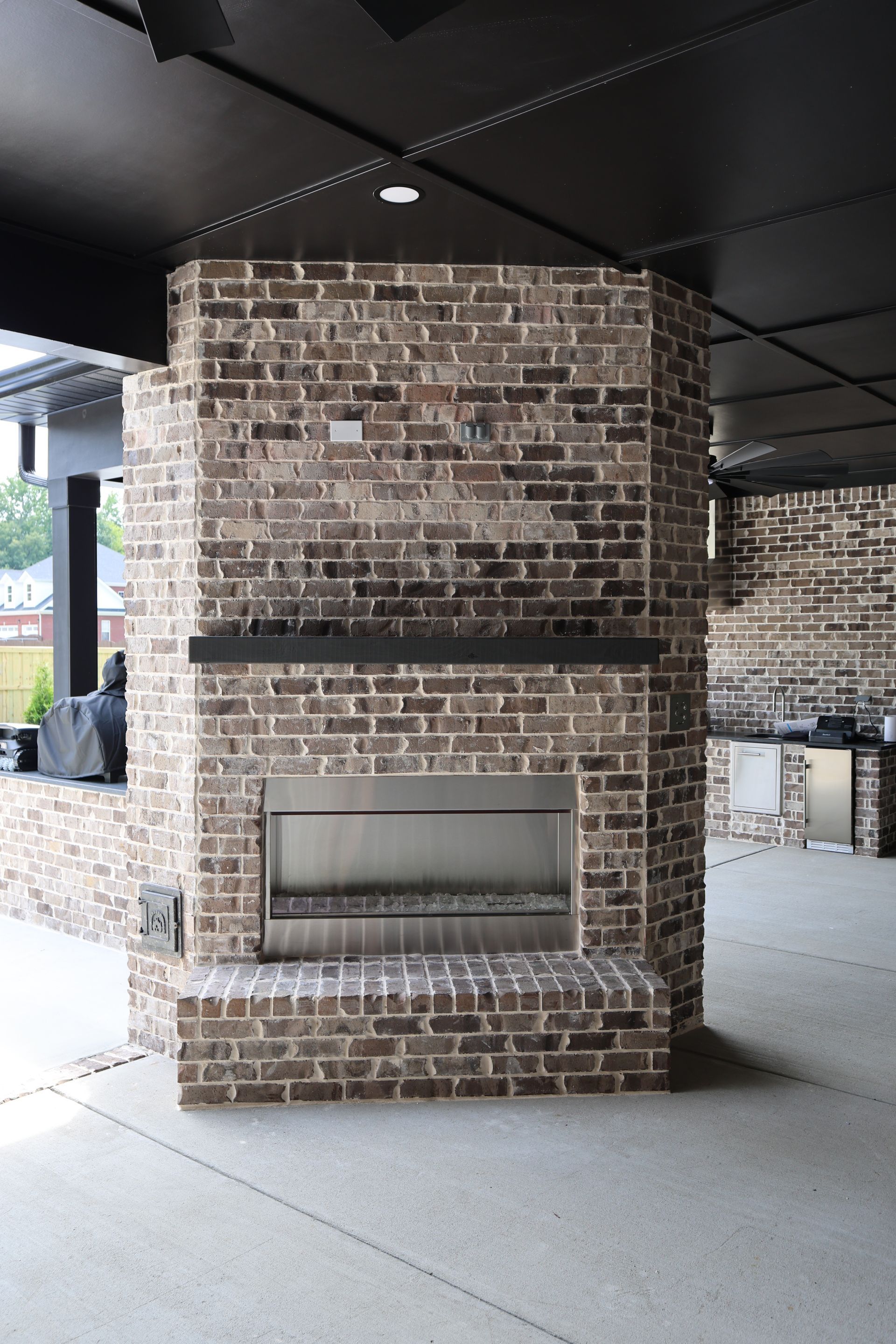 Brick outdoor fireplace under a black covered patio with stainless steel accents.