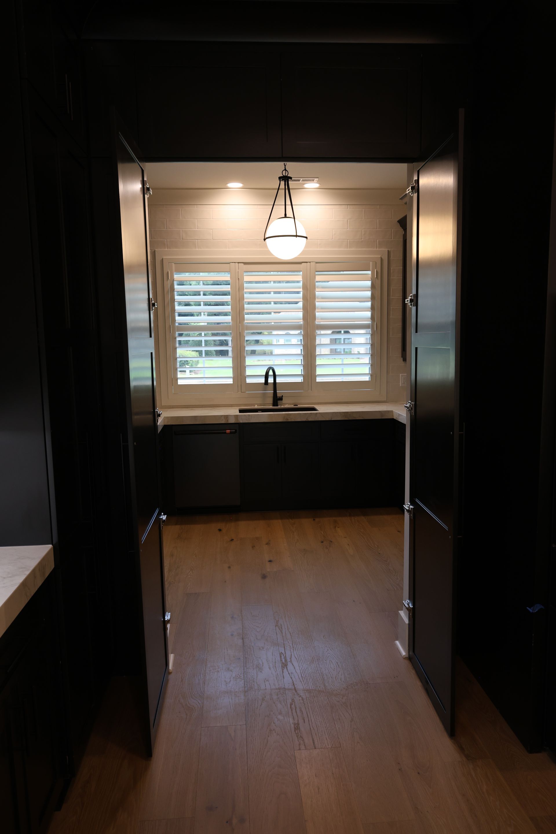 View through dark doorway into a kitchen with dark cabinetry, window with shutters, and wood floors.