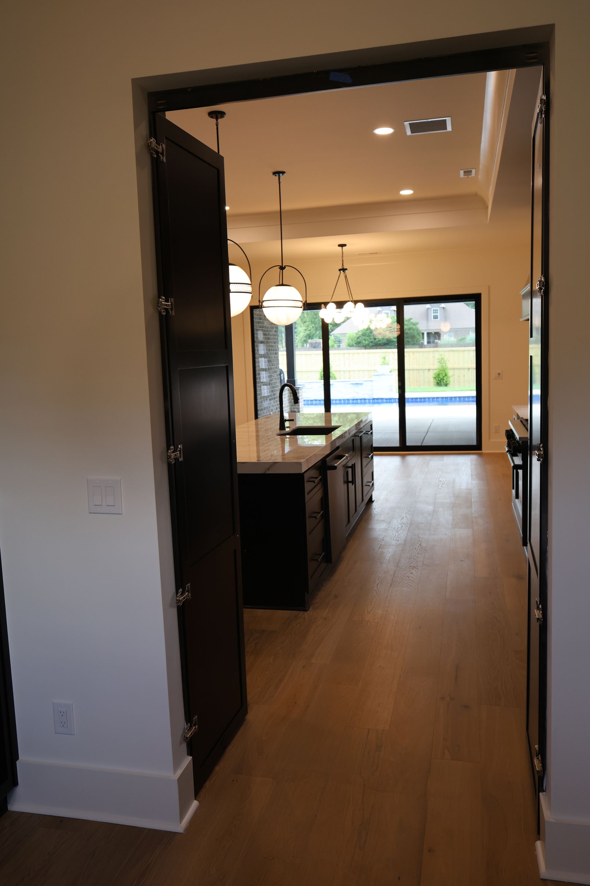 Open doorway revealing a kitchen with a dark island, pendant lights, and pool view through a glass door.