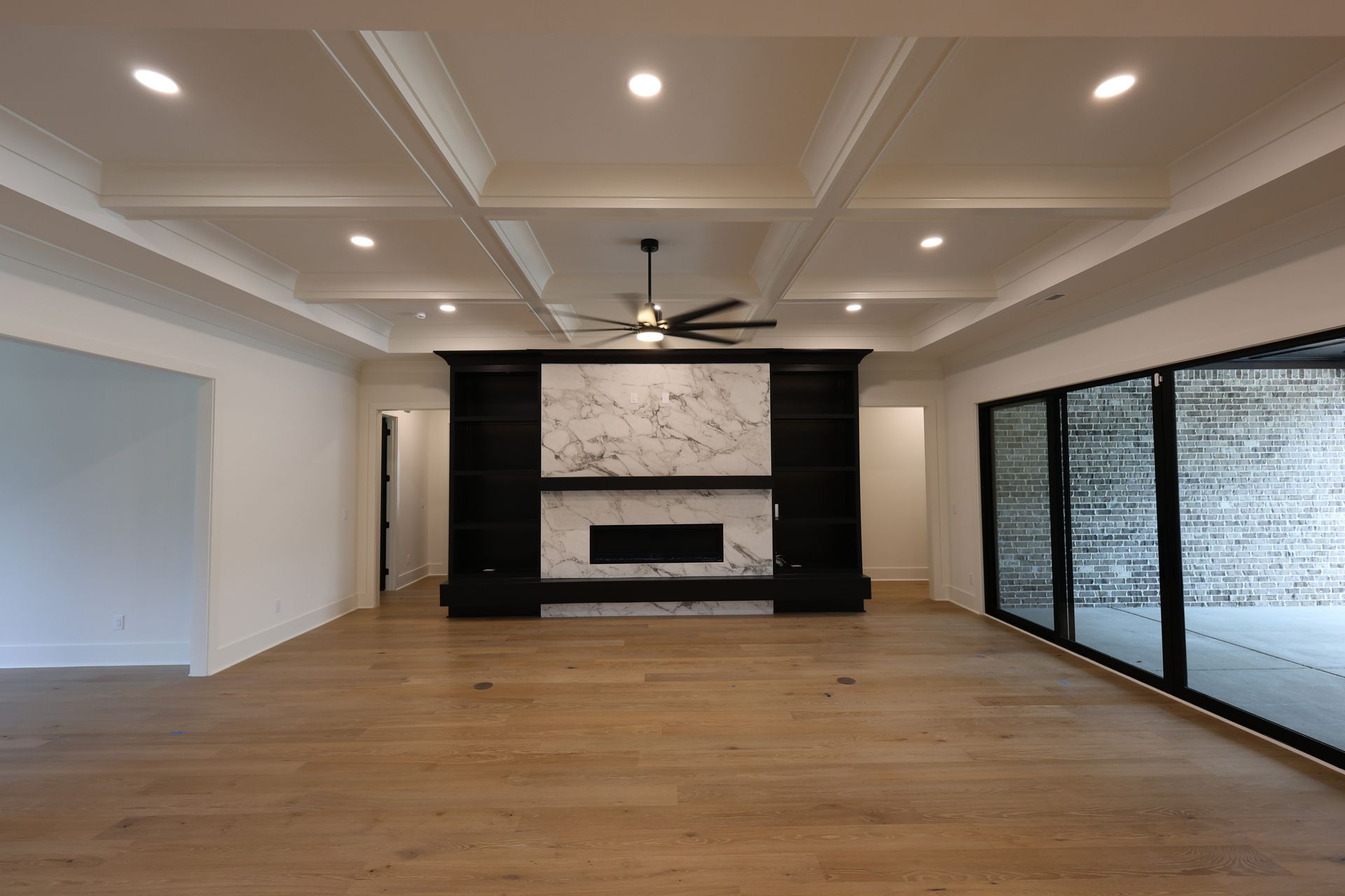 Spacious living room with fireplace, built-ins, and glass doors to a patio. Light wood floors, white walls, coffered ceiling.