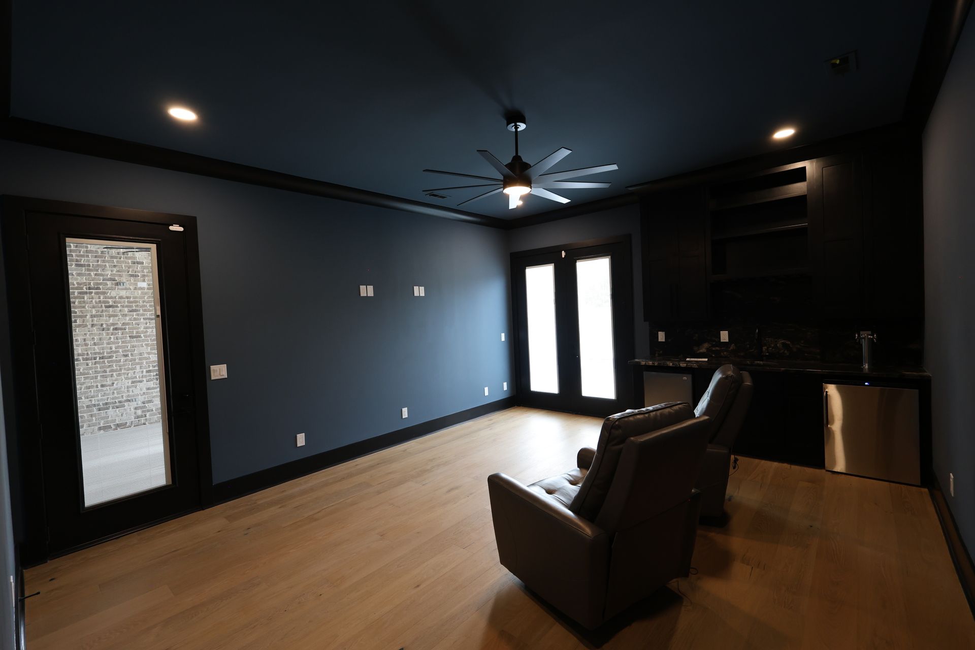 Empty dark blue media room with light wood floor. Two chairs face glass doors and small bar.