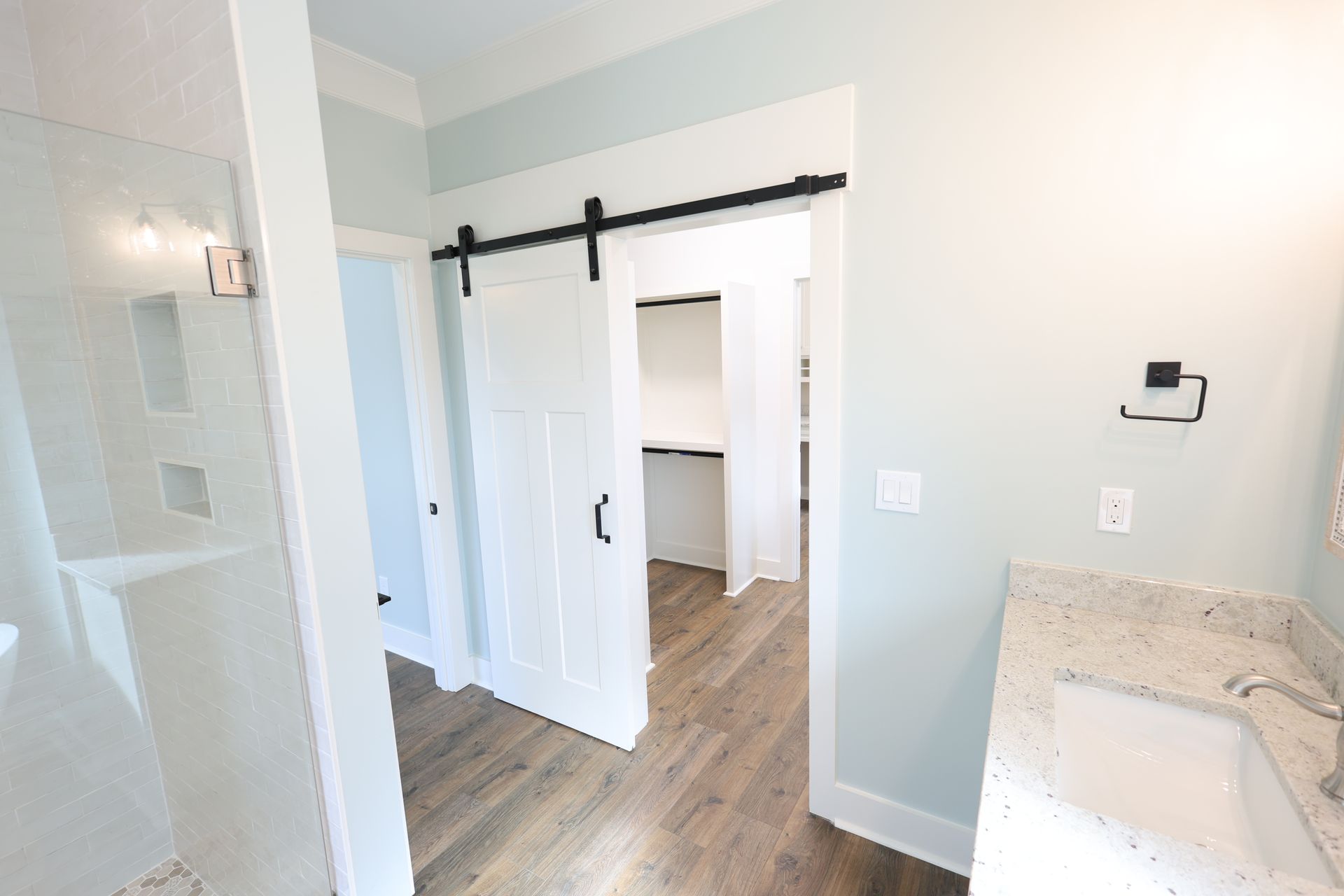 Bathroom interior with a white door, shower, and vanity. Light blue walls and wooden floor.