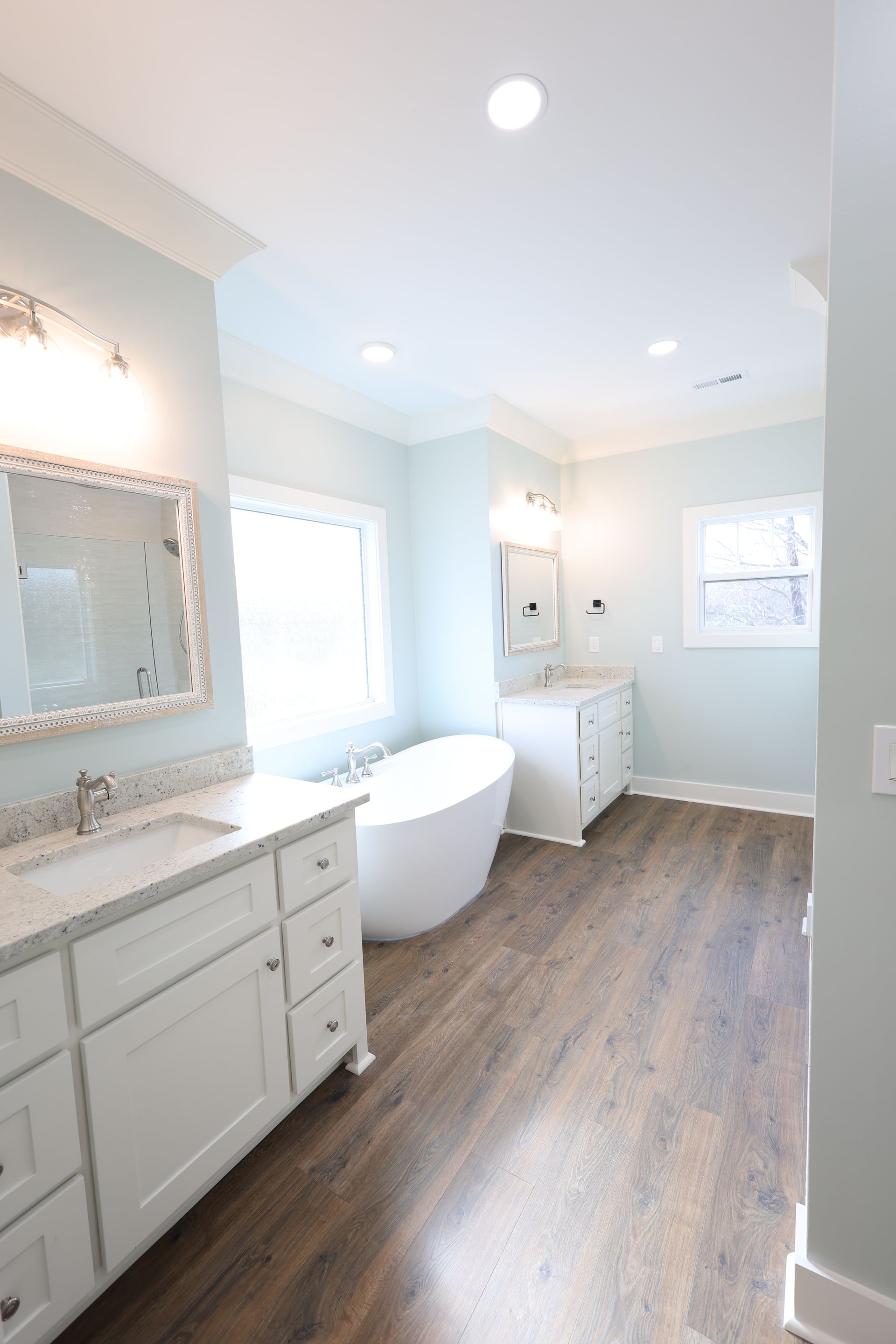 Spacious, light blue bathroom with white cabinets, freestanding tub, and wood-look flooring.