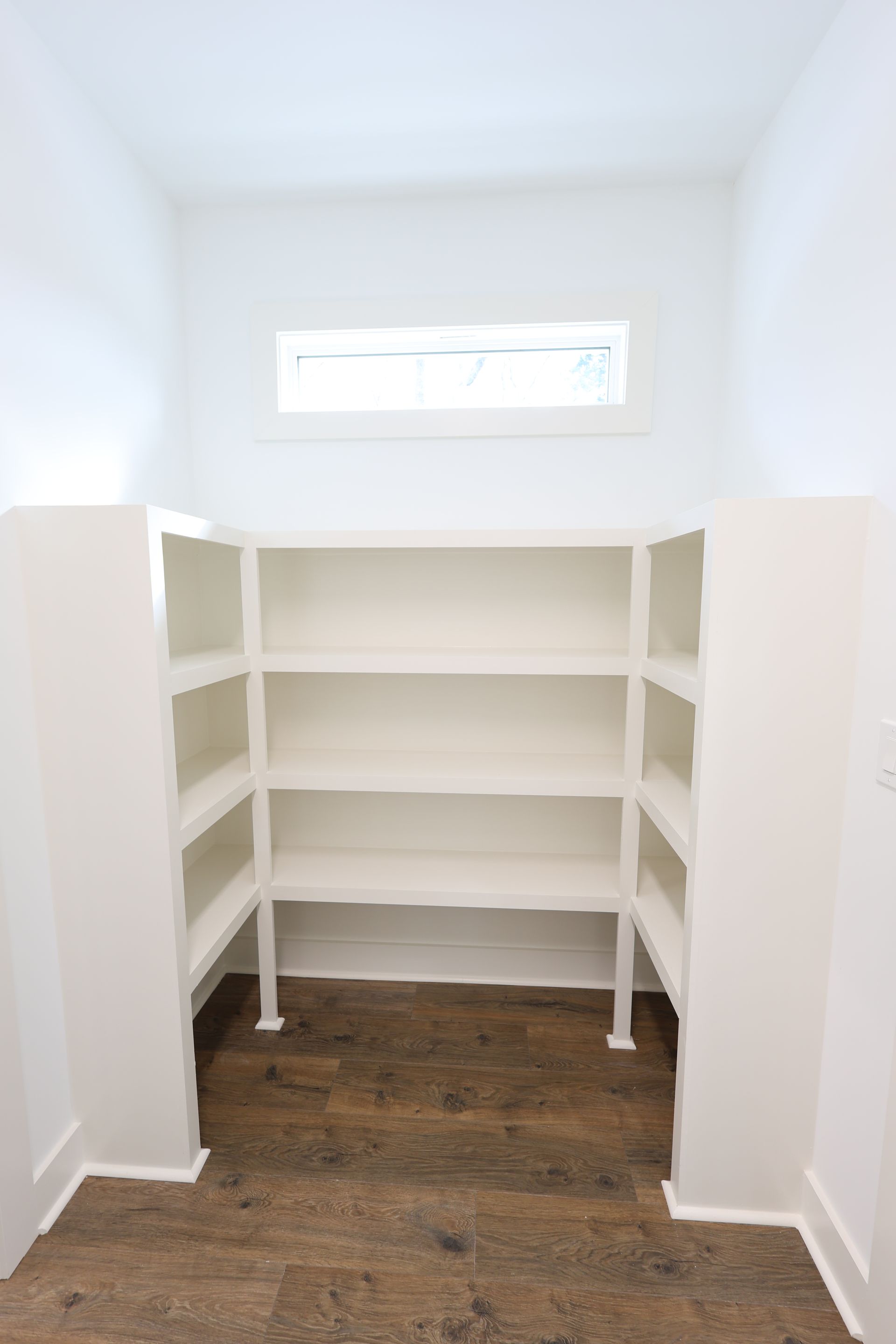 White shelving unit in a pantry with a window, against white walls and wood floor.