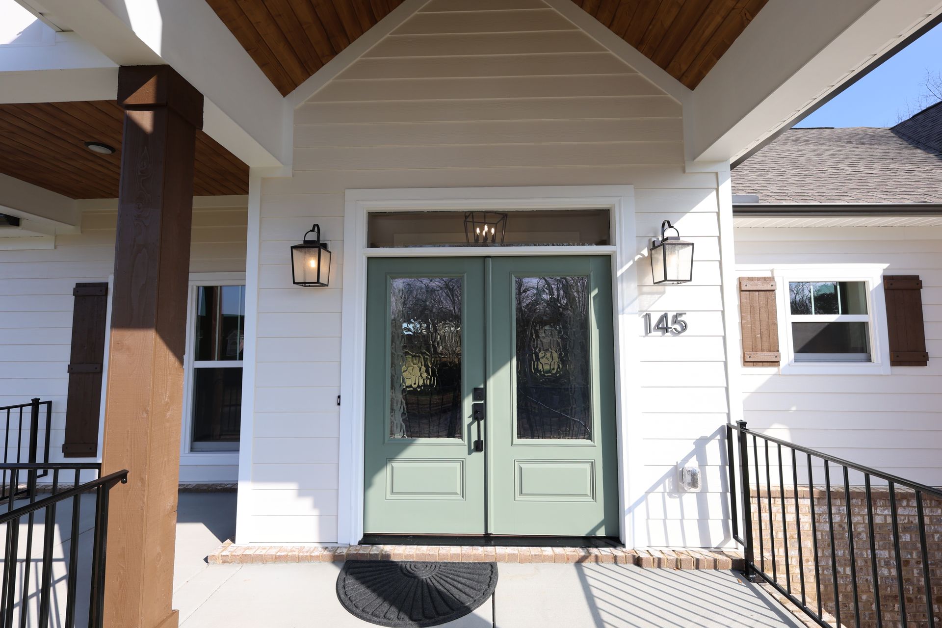 A light green double door with glass panels on a white house, flanked by brown shutters and sconces.