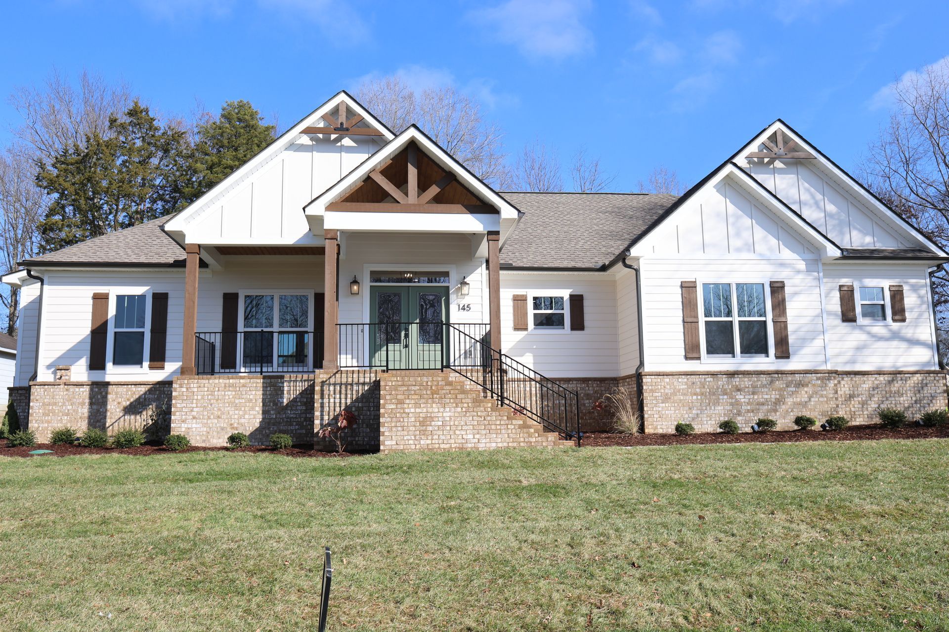 White house with brown trim and shutters, green door, and stone base against a blue sky.