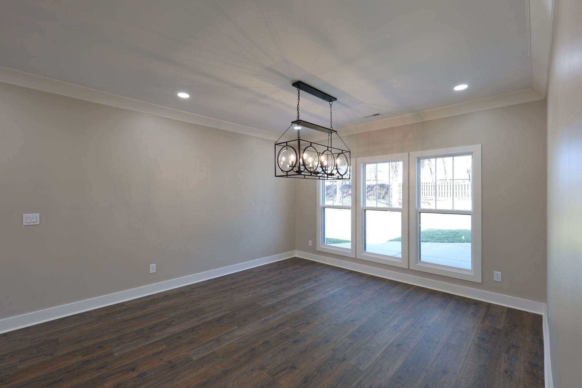 Empty dining room with dark wood floor, windows, and a black chandelier. Beige walls, white trim.