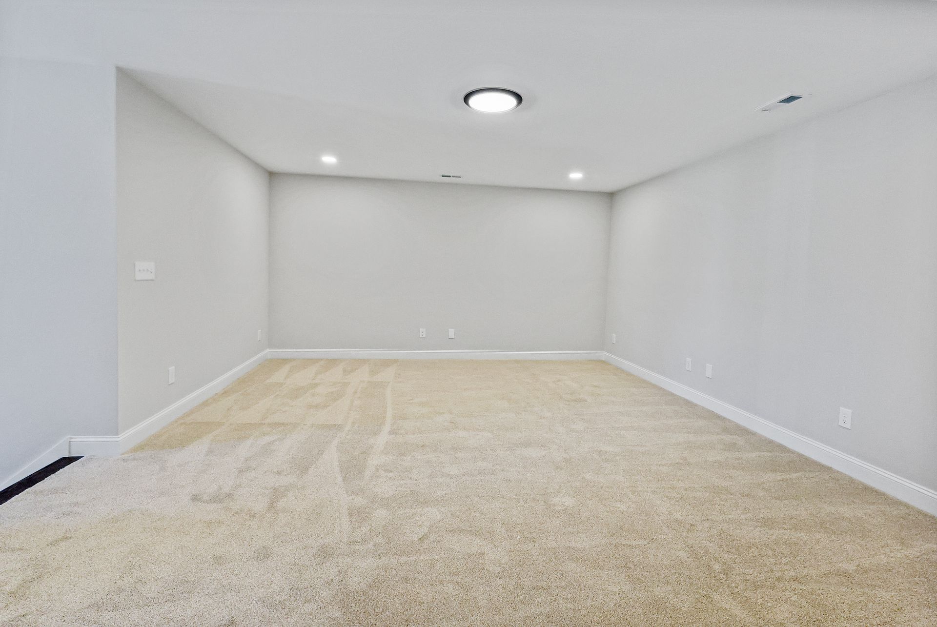Empty room with beige carpet and light gray walls, lit by recessed and ceiling lights.