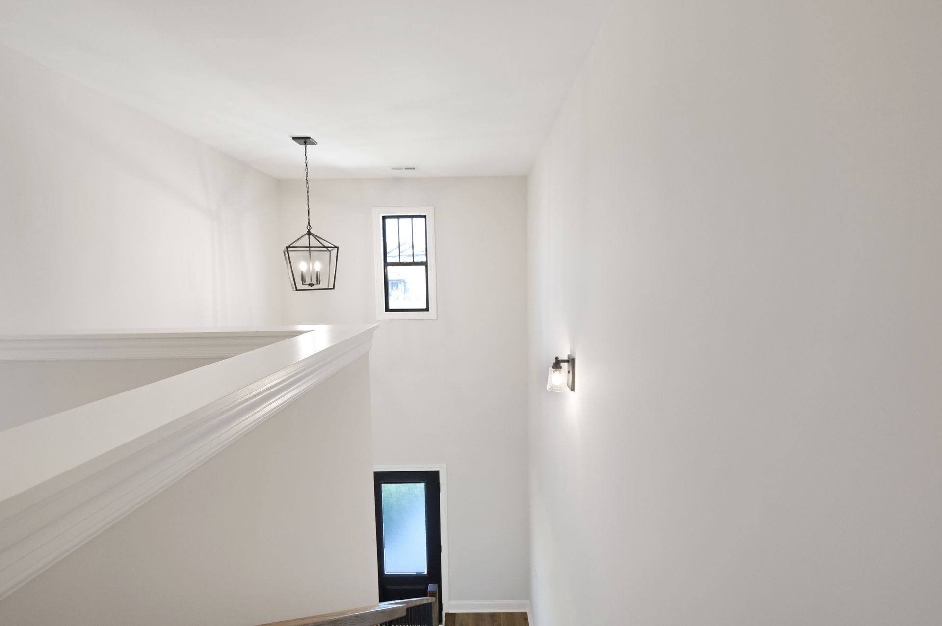 White hallway with staircase, pendant light, small window, black door, and wall sconce.