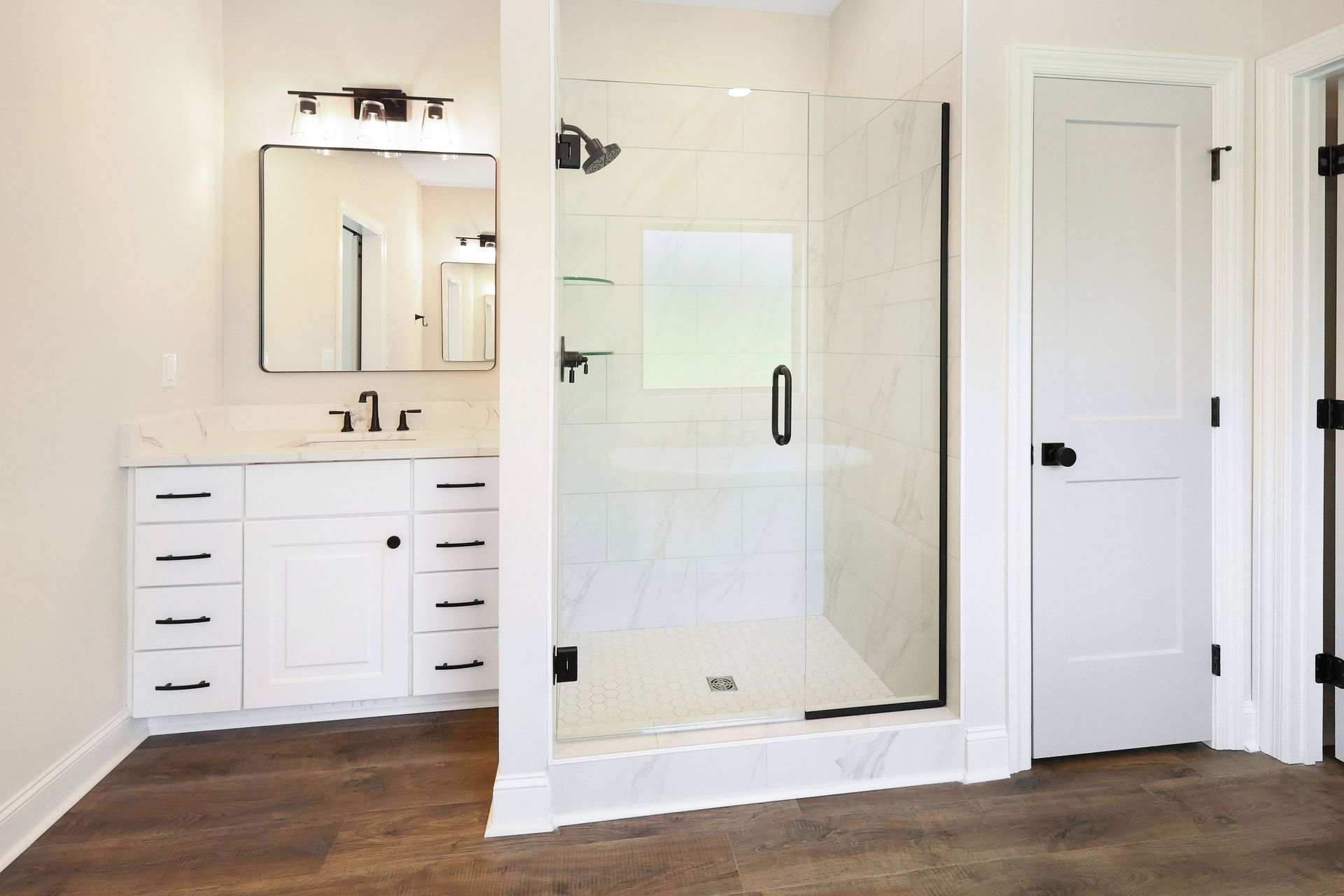 White bathroom with a glass shower, vanity, and closed door, all with black hardware, on brown flooring.