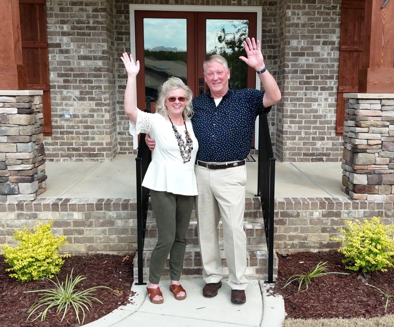 Couple waving in front of a brick house with a stone porch. They smile, and it's daytime.