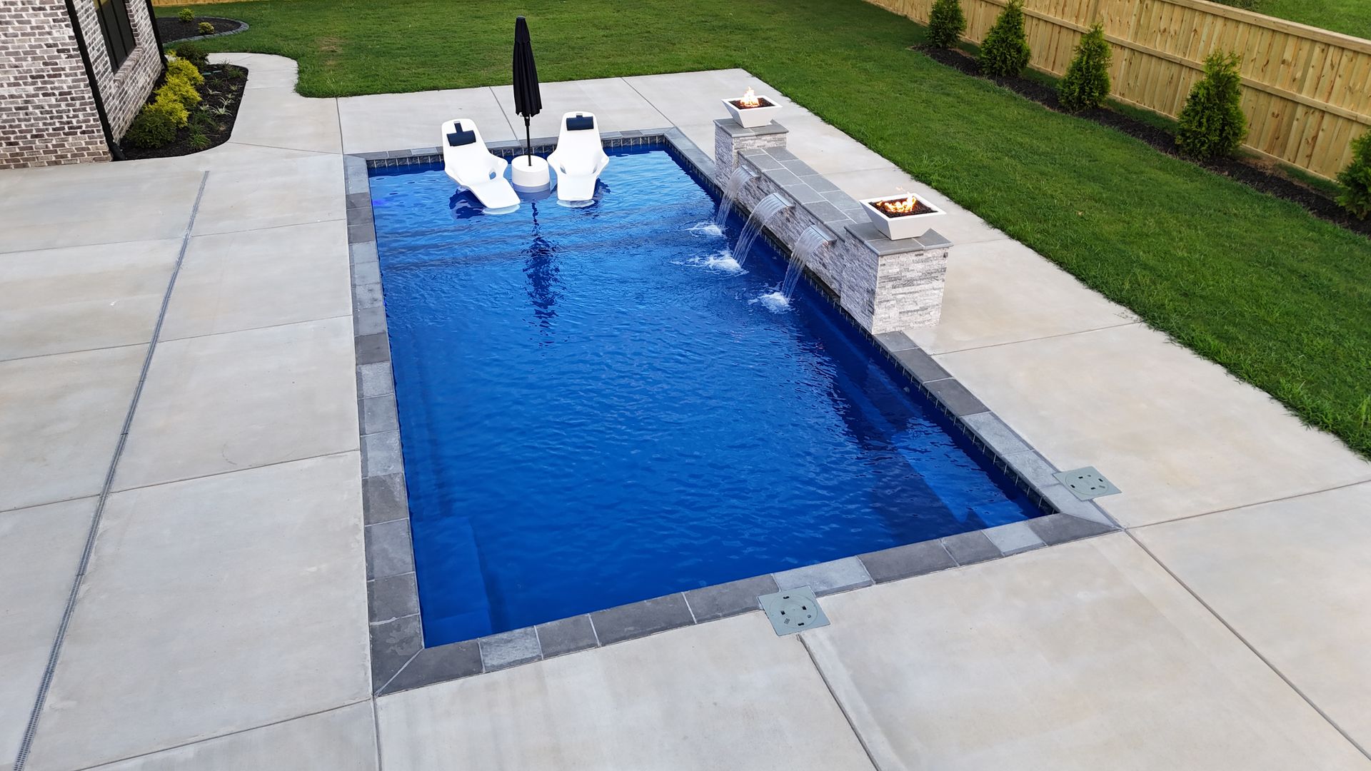 Rectangular blue pool with stone surround, concrete patio, small waterfall, and green lawn.