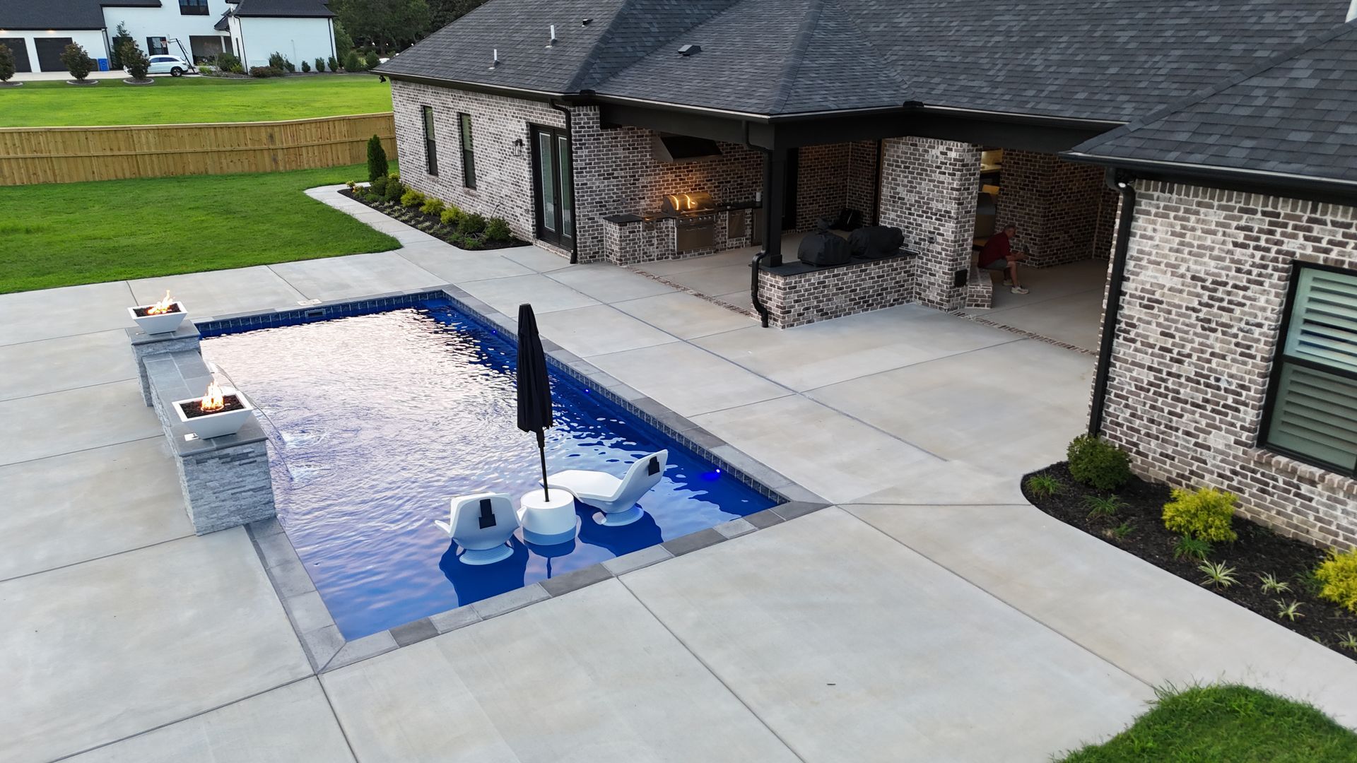 Backyard with rectangular pool, patio, and house. Pool has fire features, floating chairs, and umbrella. Brick exterior.