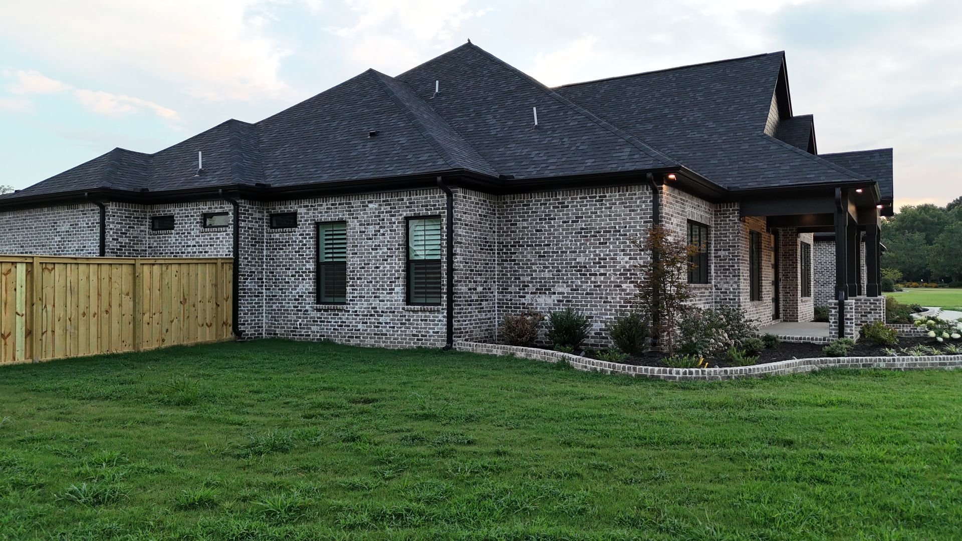 House with gray brick exterior, dark roof, wooden porch, and fenced yard on a grassy lawn.