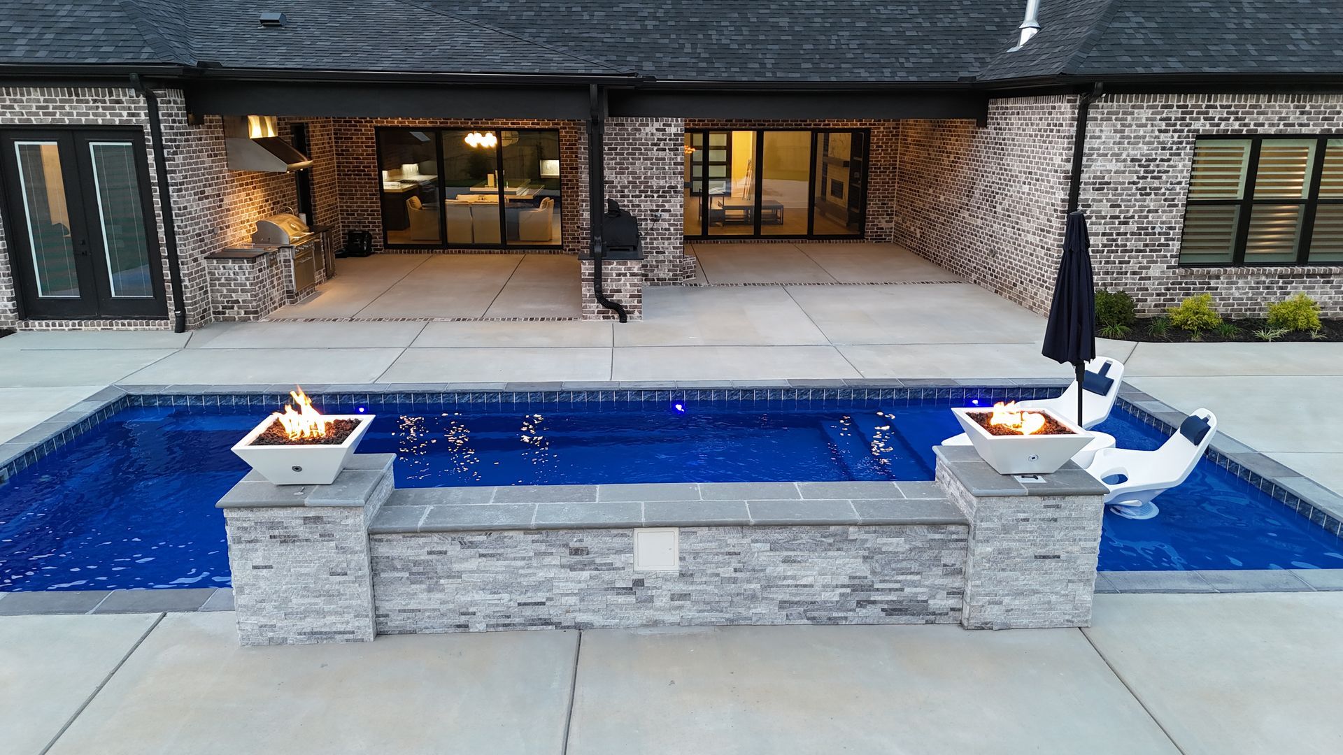 A backyard pool with fire features, brick house exterior, patio furniture, and dark blue water.