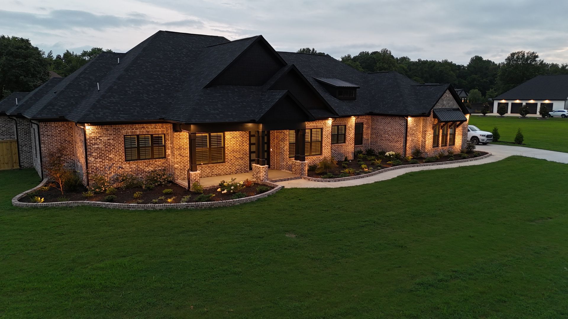 Brick house at dusk, lit by exterior lights, with a green lawn and trees in the background.