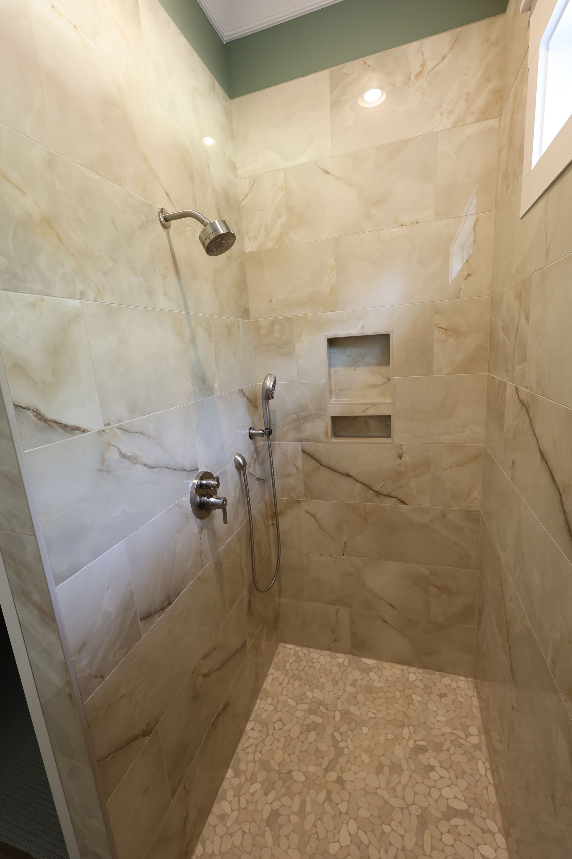 Tiled shower with handheld and overhead showerheads. Two recessed shelves and light-colored marble effect tiles.