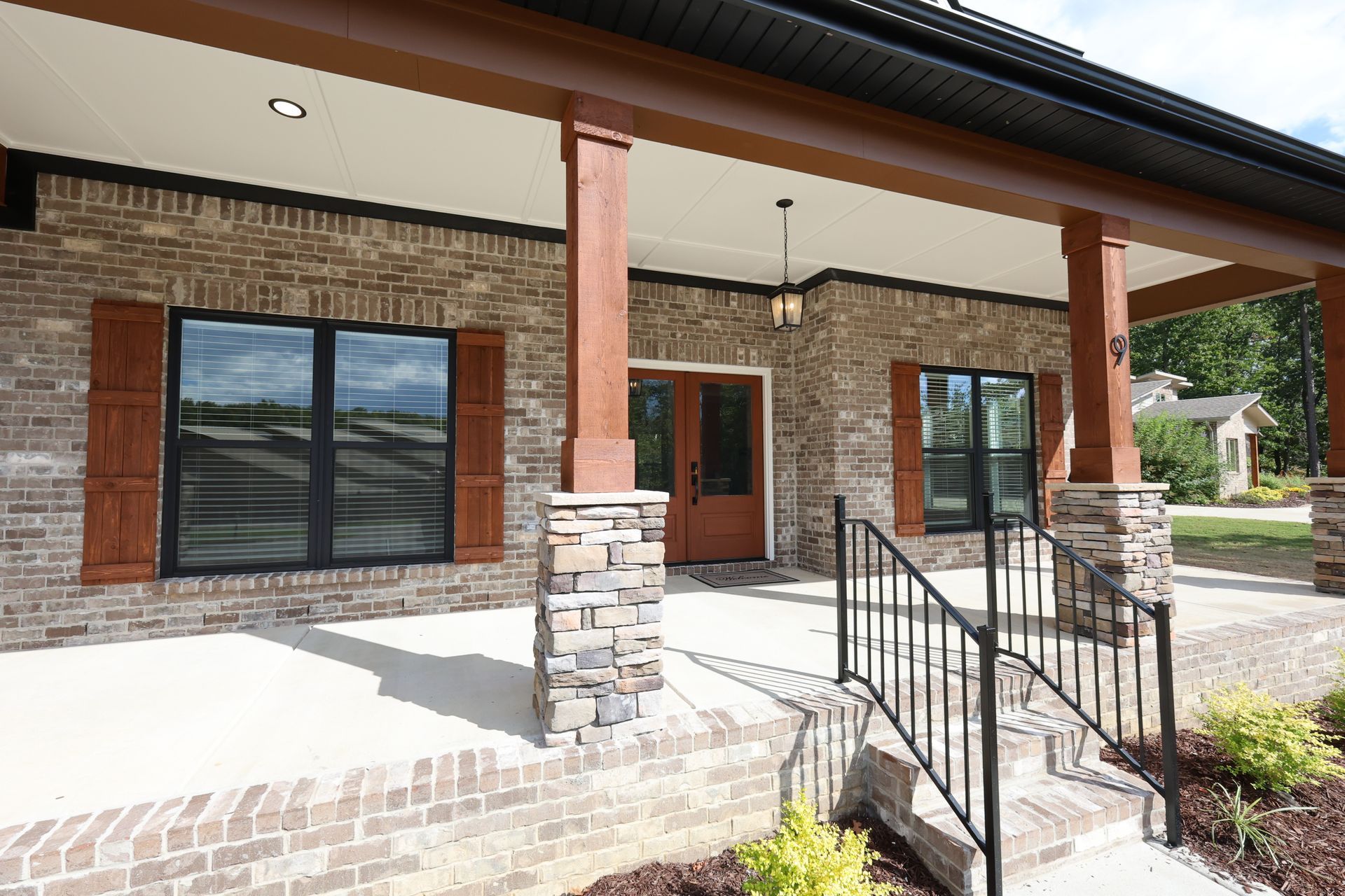 Brick house with covered porch, wooden beams, stone columns, black windows, and brown shutters.