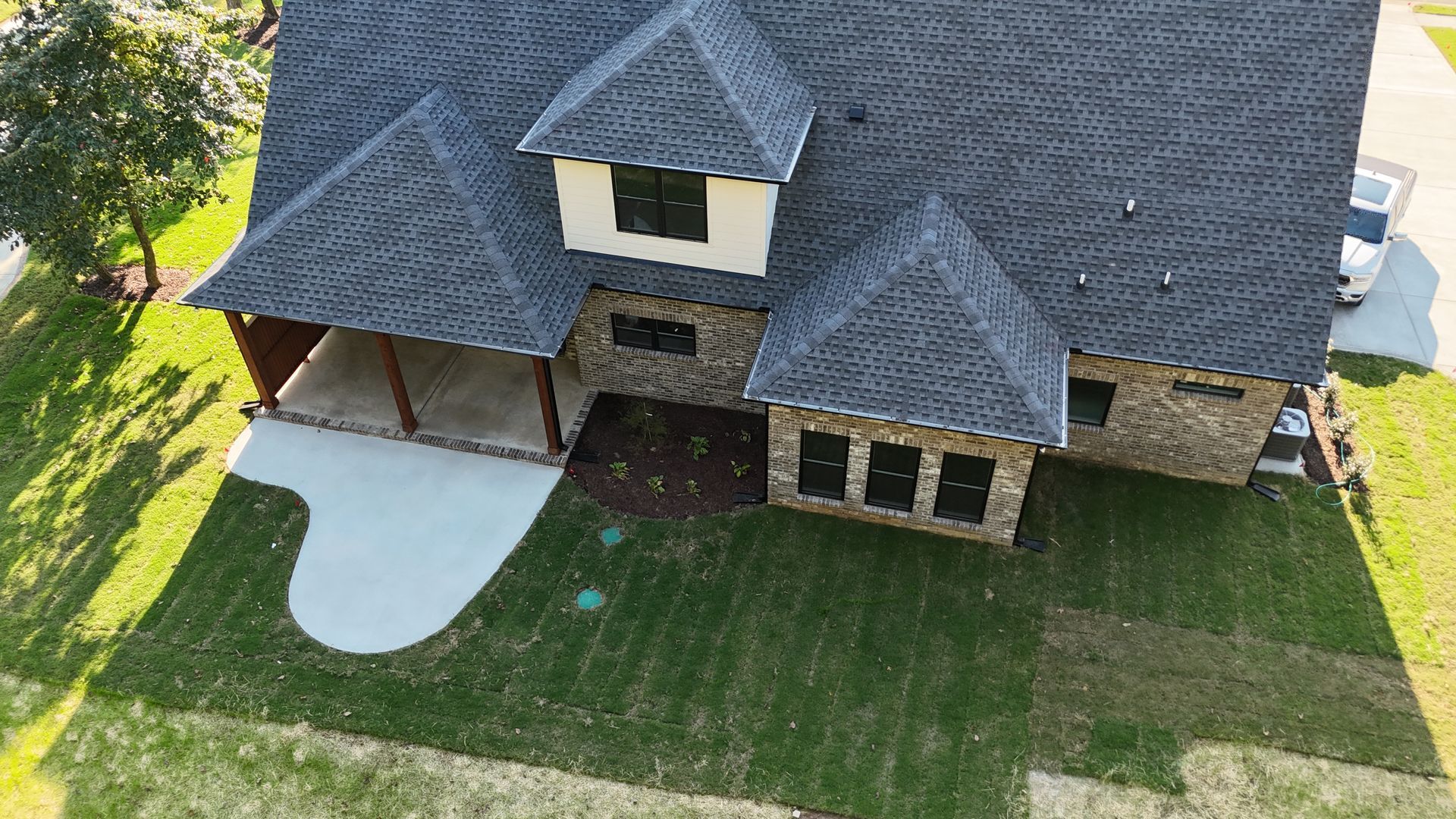 Aerial view of a house with a stone facade and dark shingle roof, green lawn, and a concrete patio.