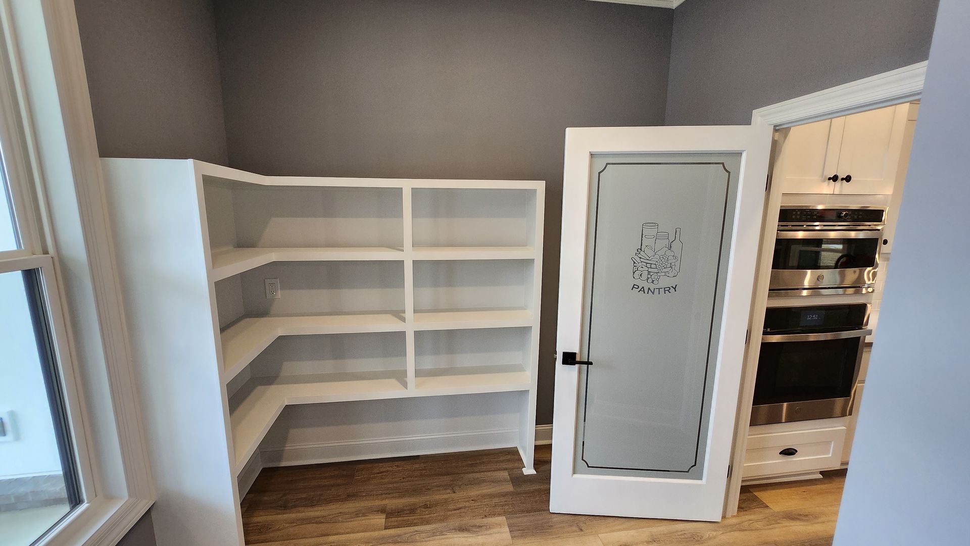 A pantry with white shelves and a frosted glass door next to built-in ovens and a window.