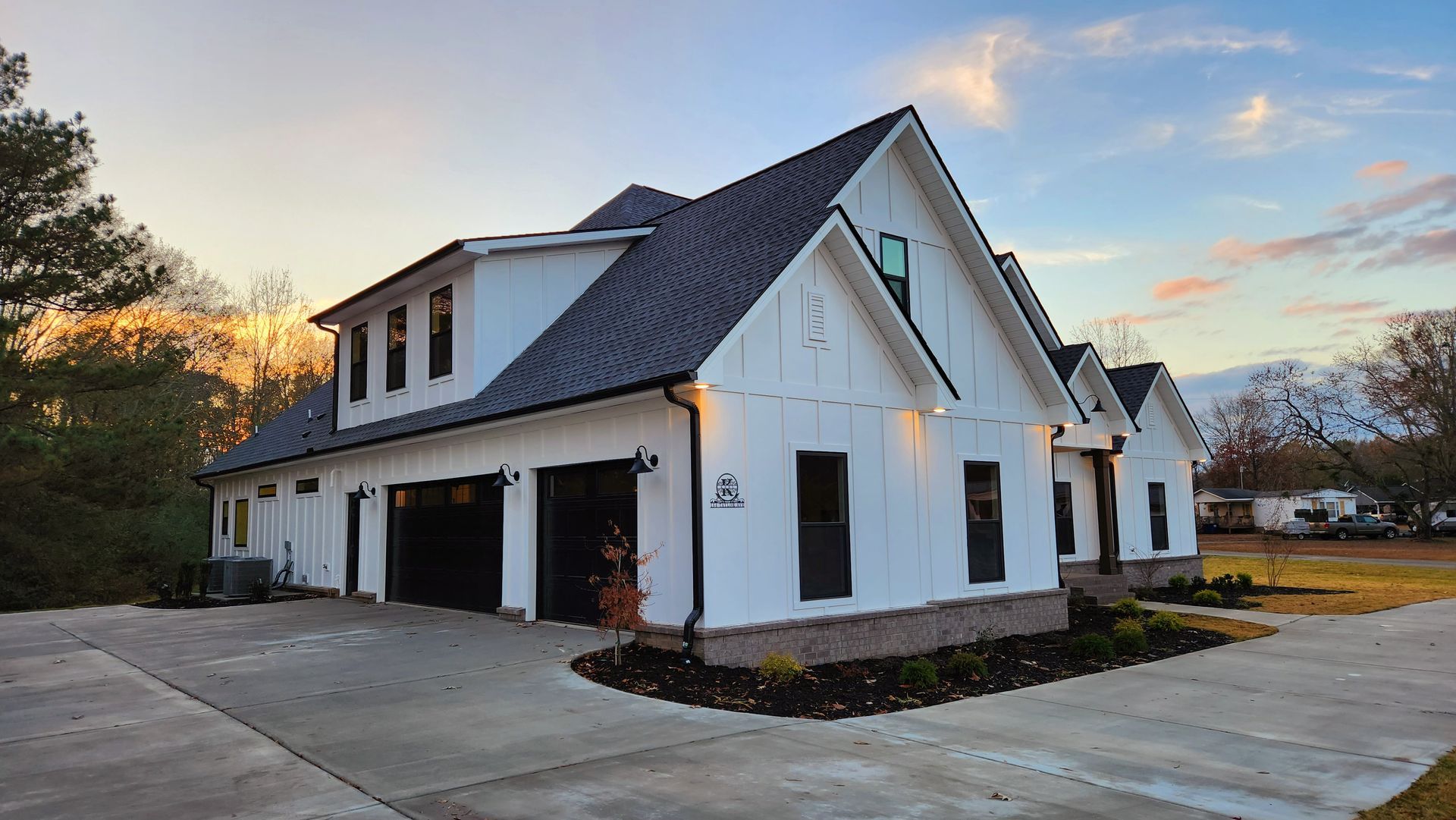 White modern farmhouse with black trim, black garage doors, gray roof, and concrete driveway against a sunset sky.