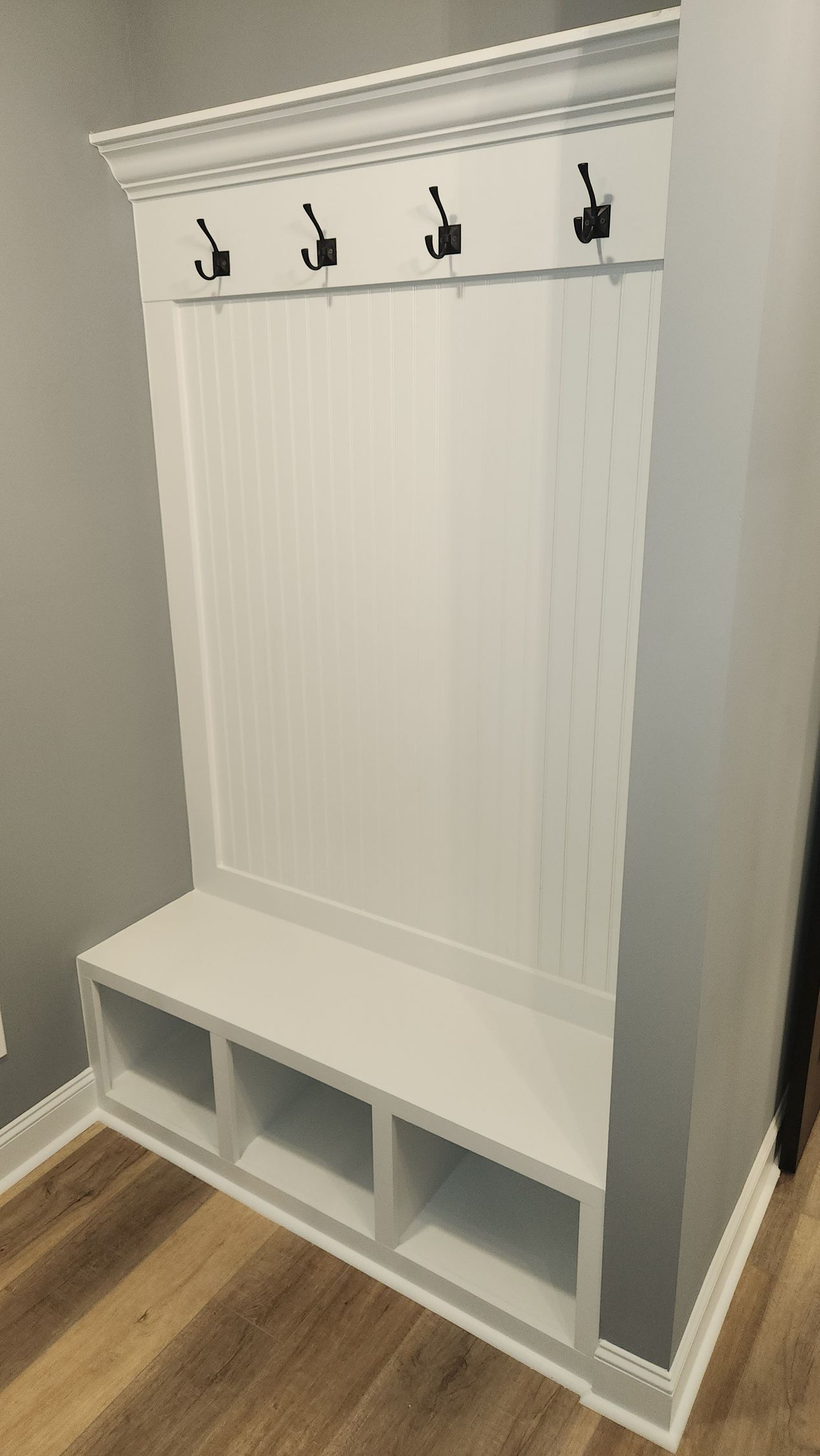 White built-in entryway bench with storage cubbies, coat hooks, and crown molding against gray walls and wood-look flooring.