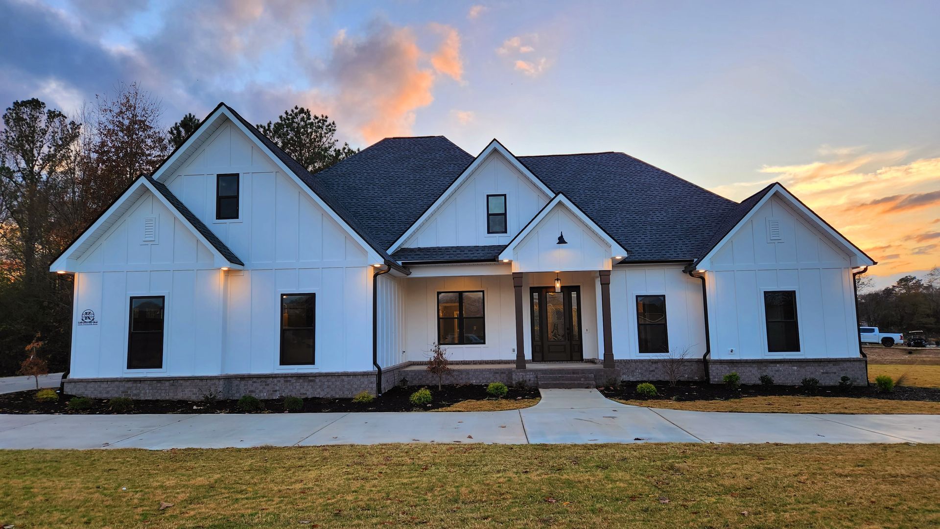 White farmhouse with dark roof, brick base, and front porch at sunset.