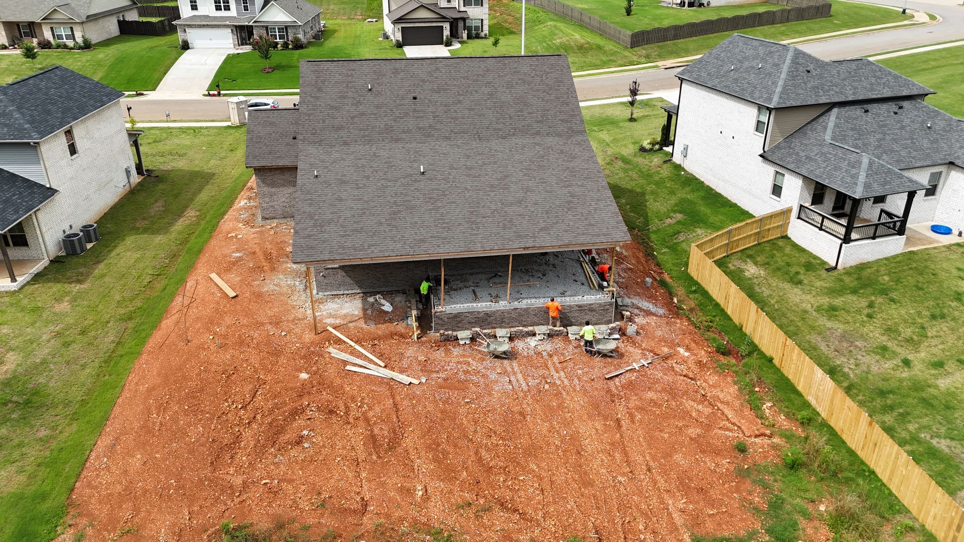 Aerial view of house under construction with workers, red dirt, and surrounding houses.