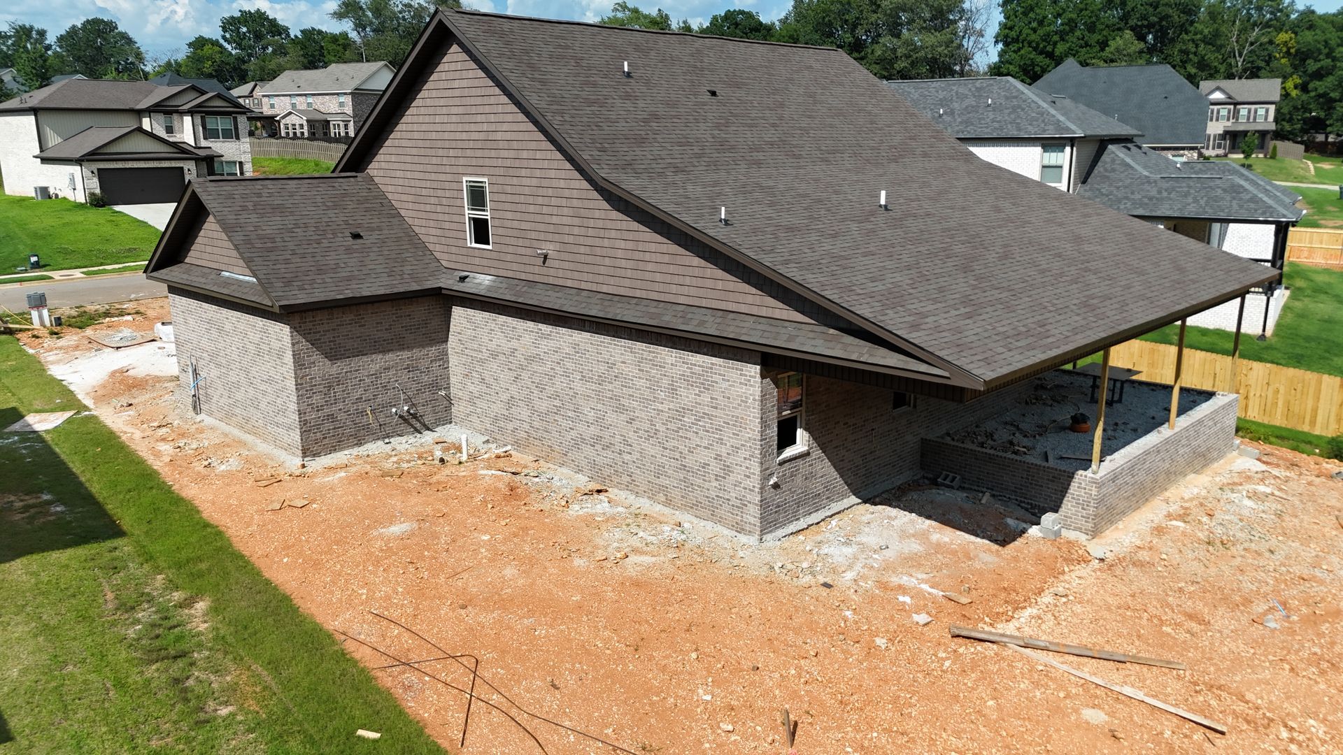 A partially constructed house with brown roof and gray brick exterior sits on a dirt lot.