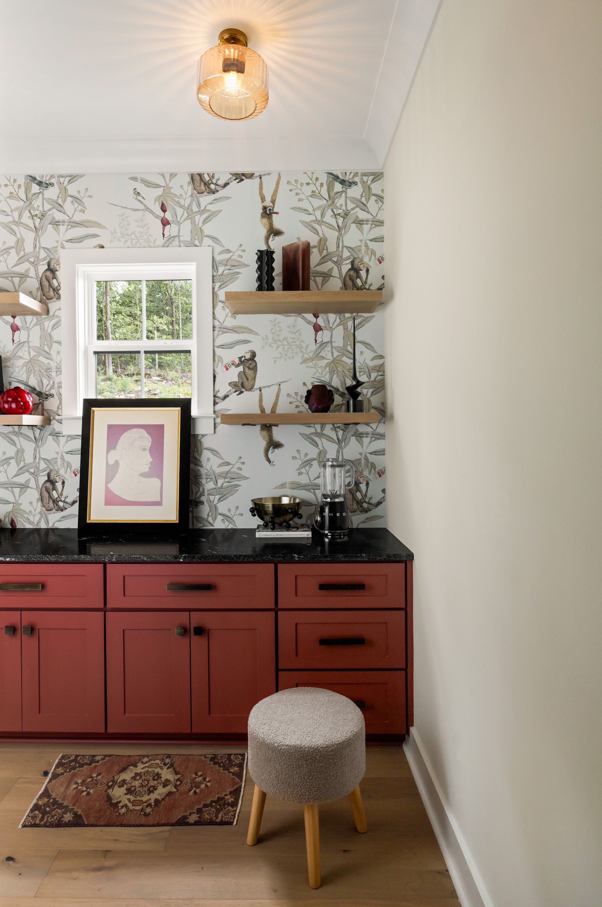 Red cabinets with black countertop, patterned wallpaper, wooden shelves, a window, and a stool.