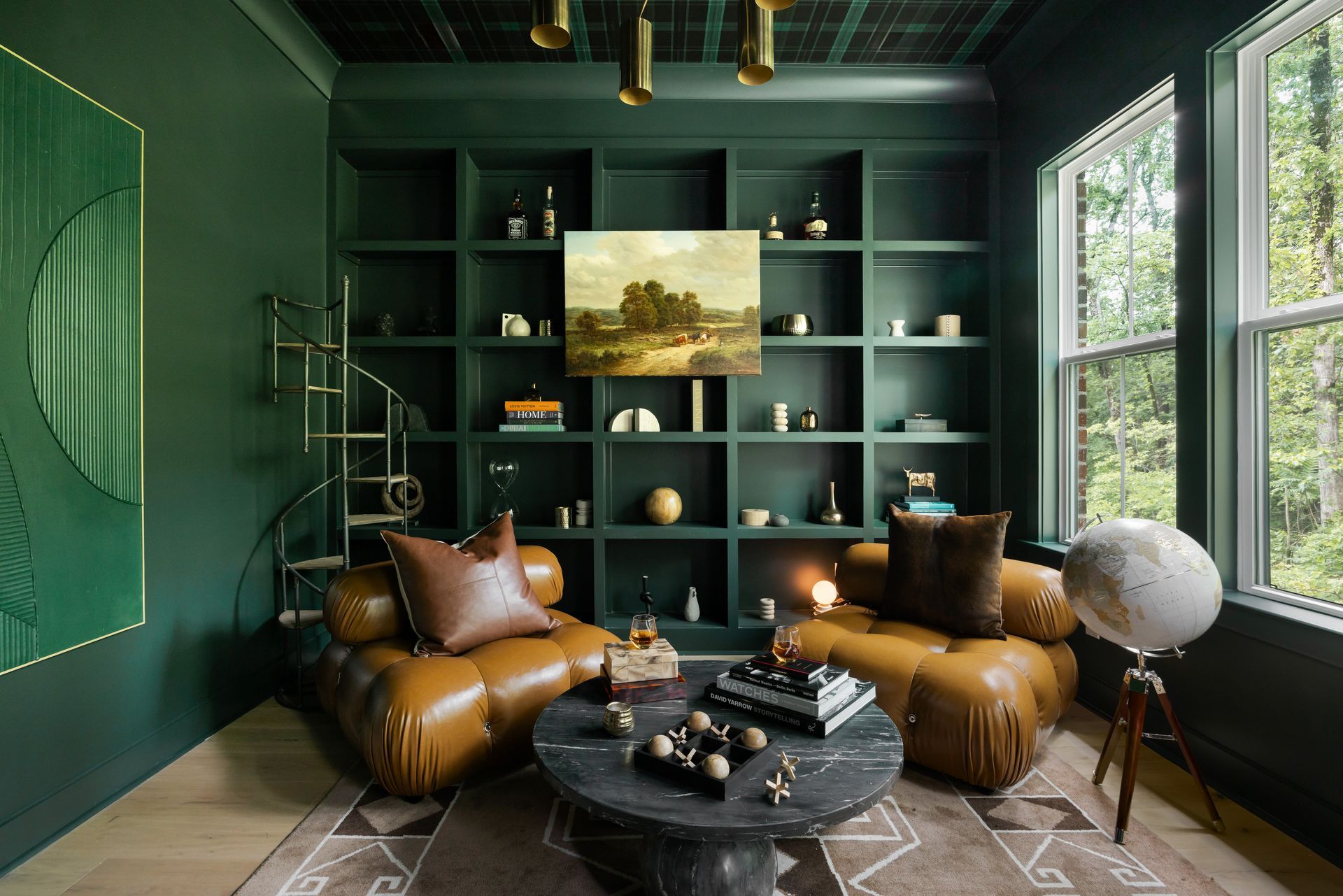 Green study with leather chairs, marble table, built-in shelving, artwork, and window overlooking trees.
