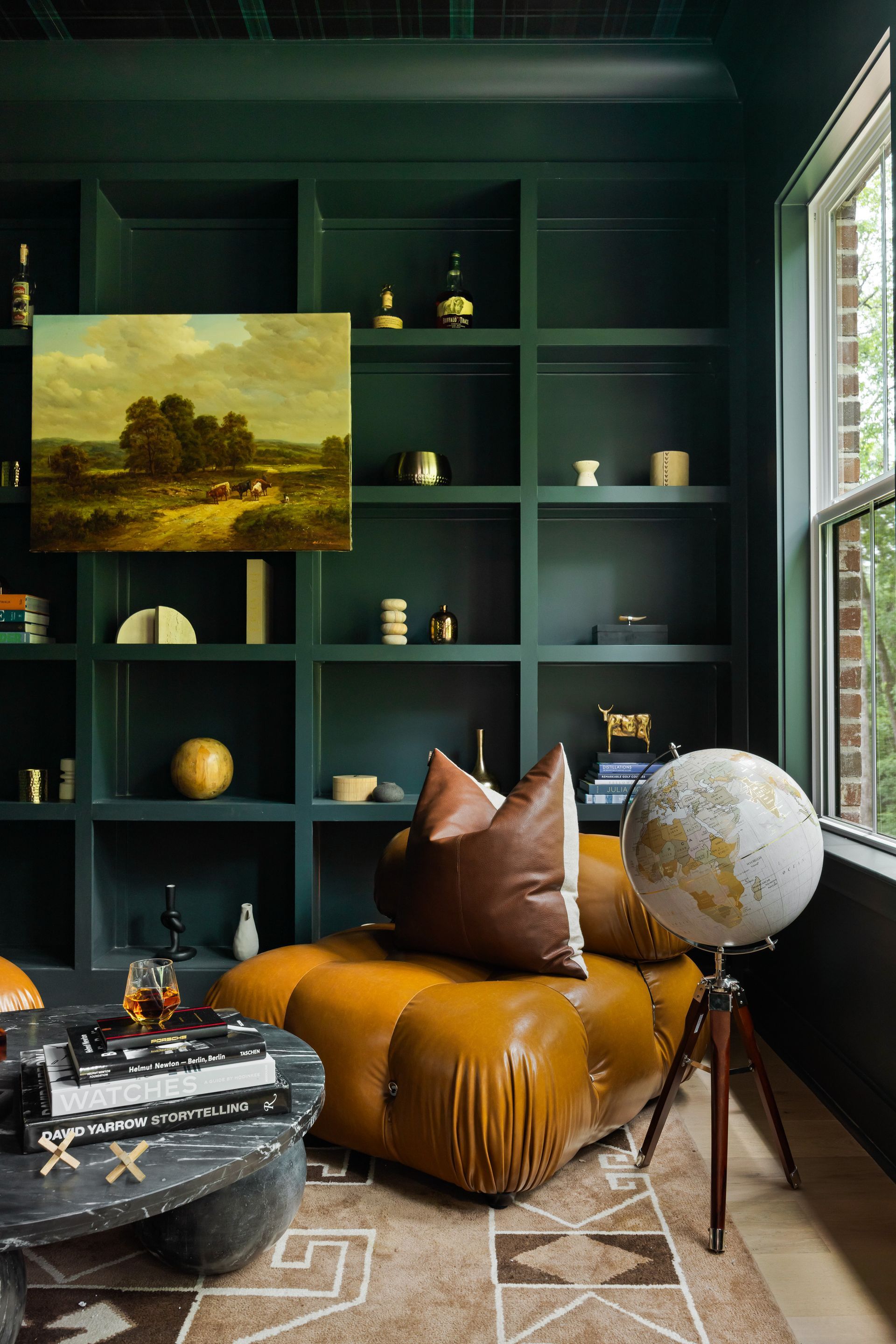 Cozy library with dark green shelves, leather armchair, marble coffee table, globe, and landscape art.