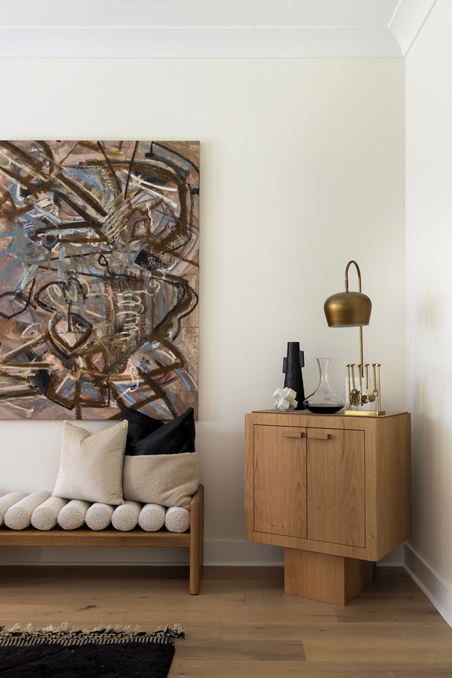 Wooden bench and cabinet with abstract art and lamp in a white-walled room.