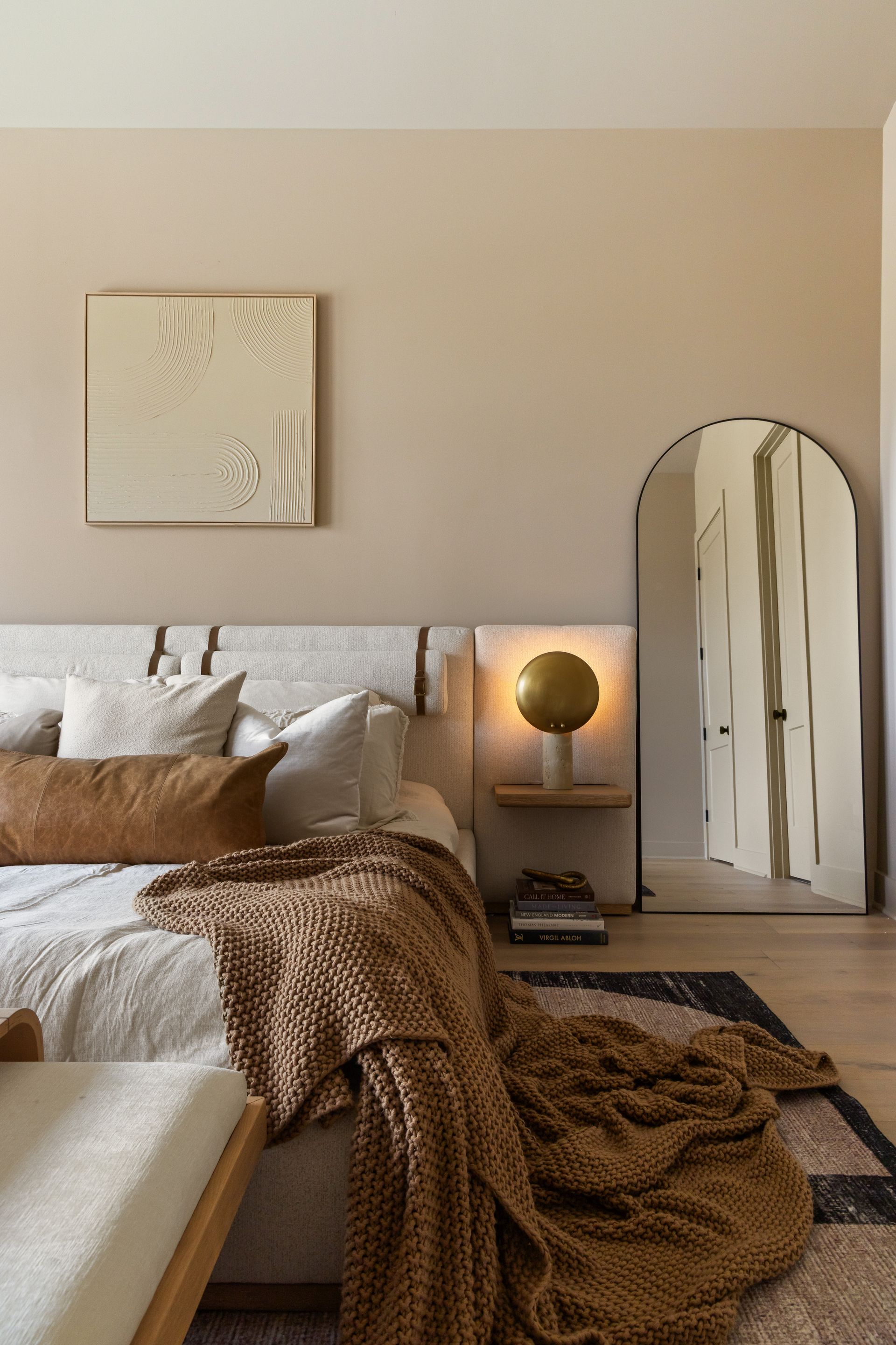 Cozy bedroom with bed, blanket, artwork, round lamp, arch mirror, and neutral tones.