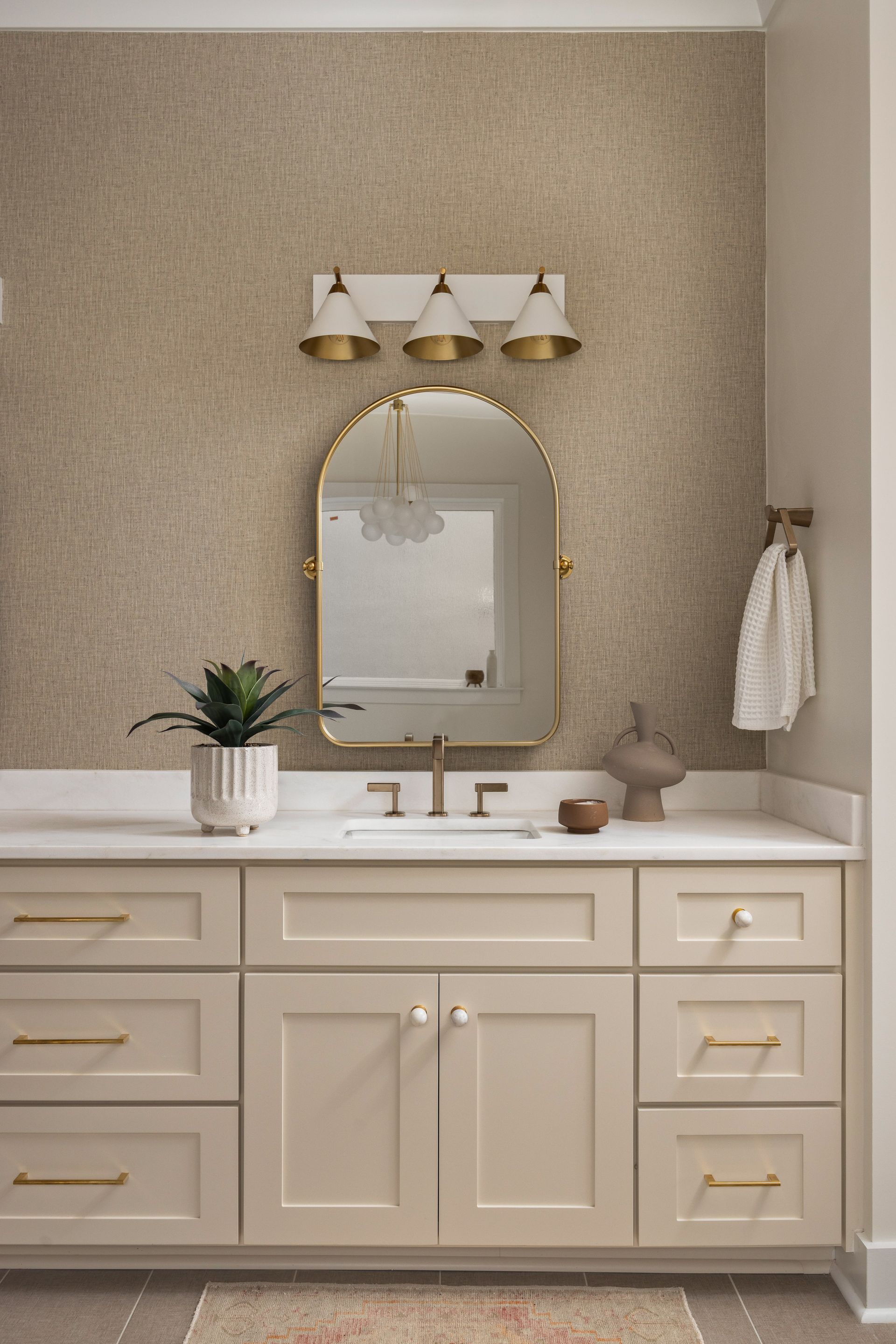 Bathroom with beige vanity, gold fixtures, textured wallpaper, arched mirror, and potted plant.