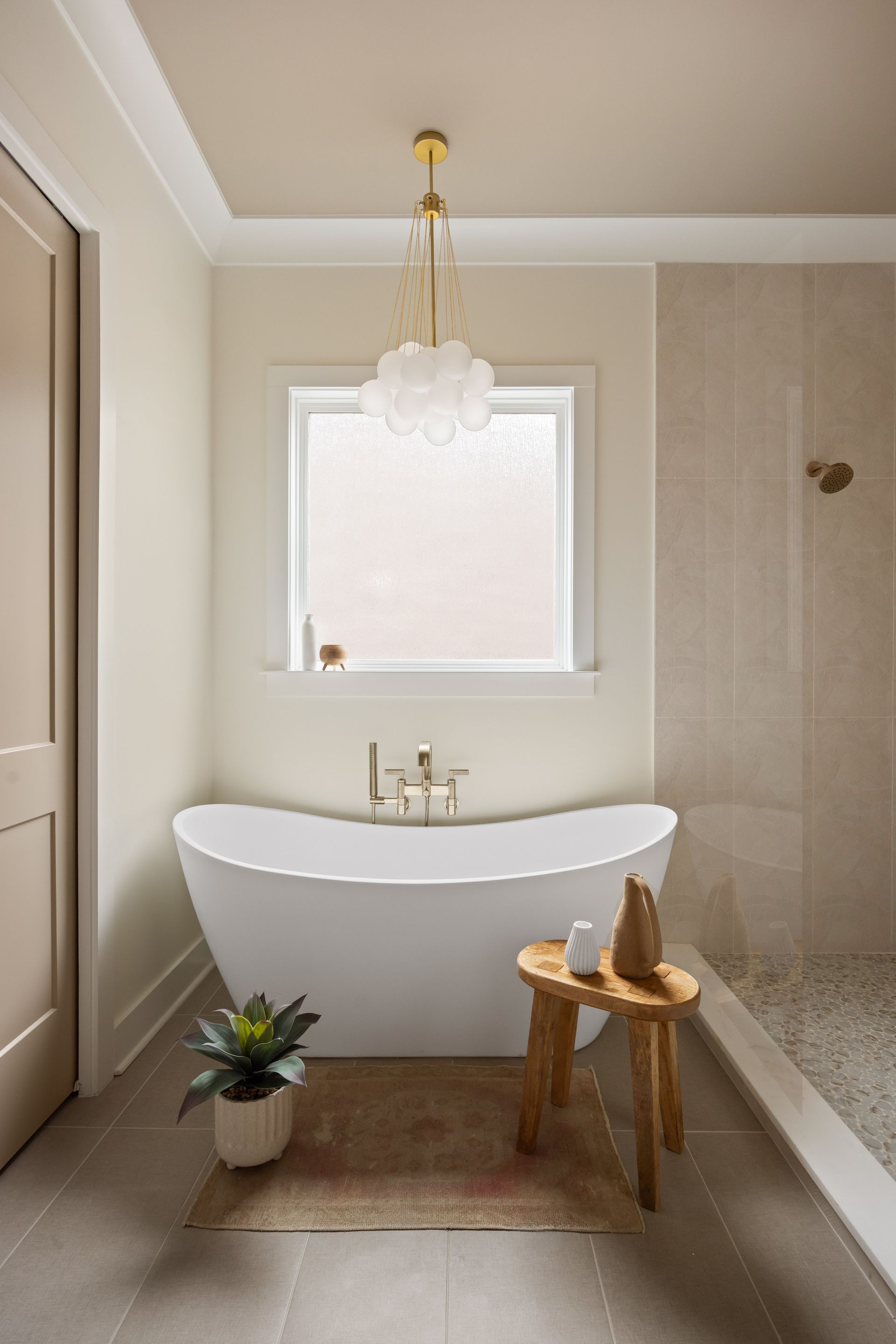 Cozy bathroom with a white tub, wooden stool, and a gold chandelier near a frosted window.