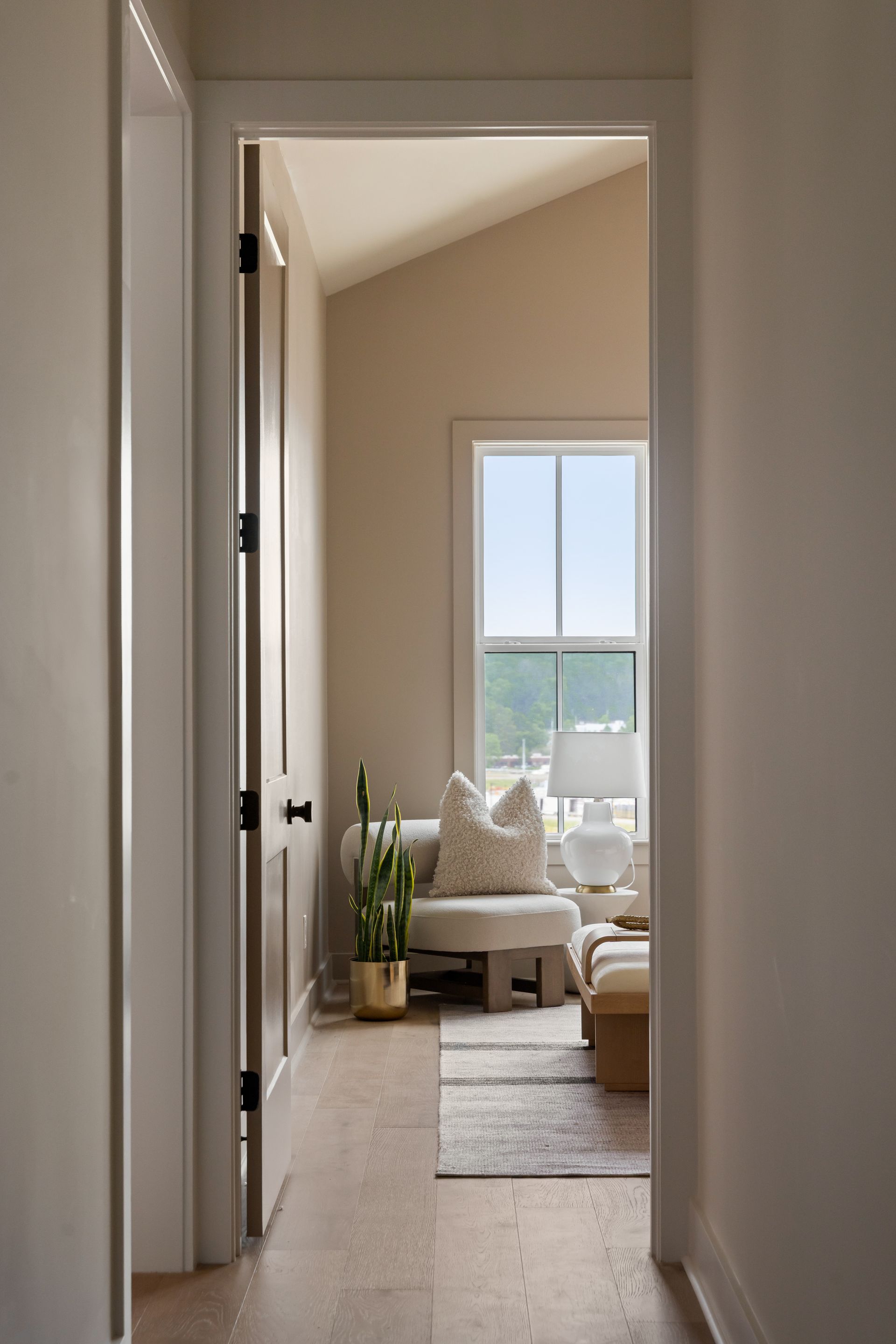 Hallway view into a room with a window, chair, and plant. Natural light and neutral colors dominate.
