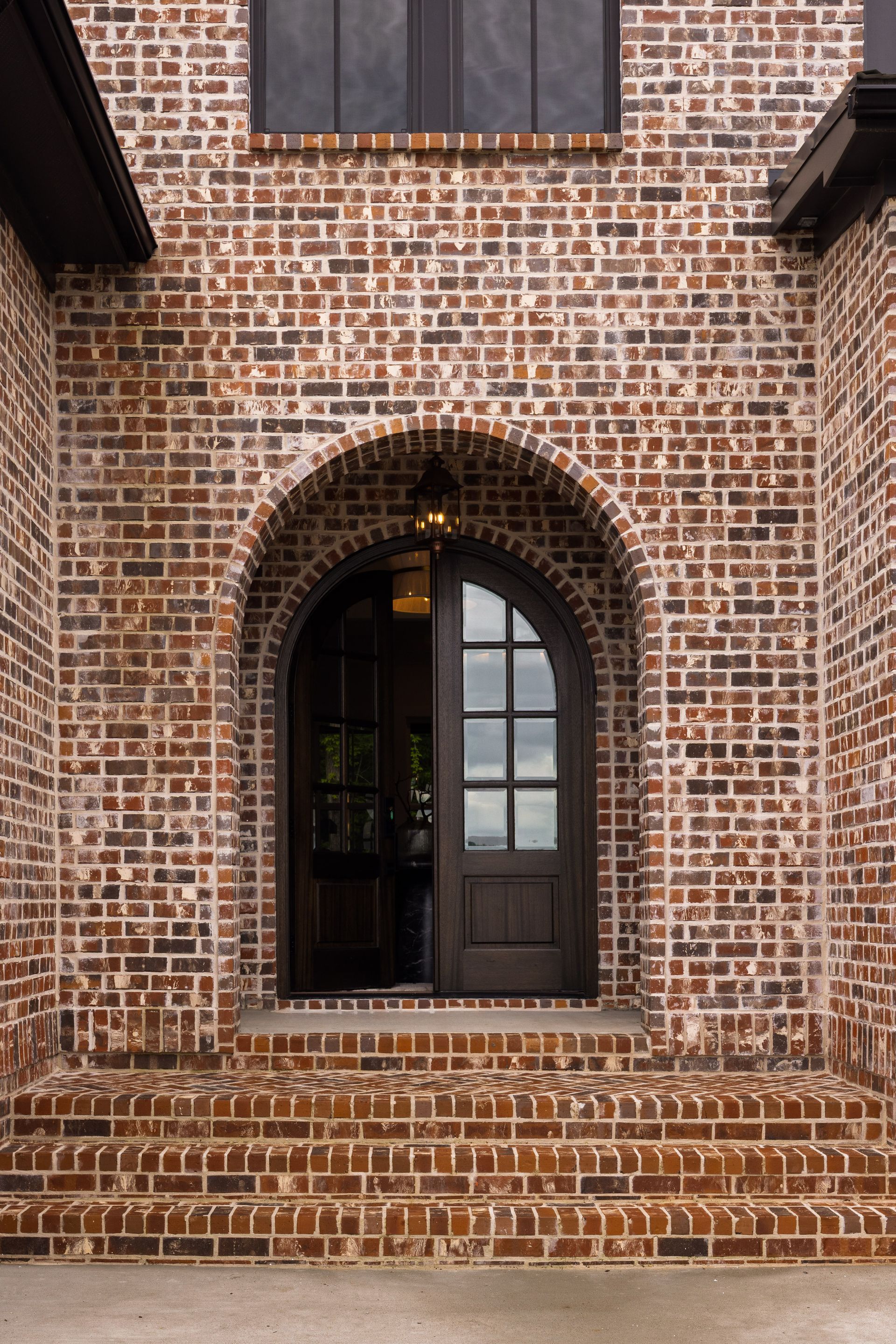 Brick arched doorway, dark wooden door, brick steps, window above.