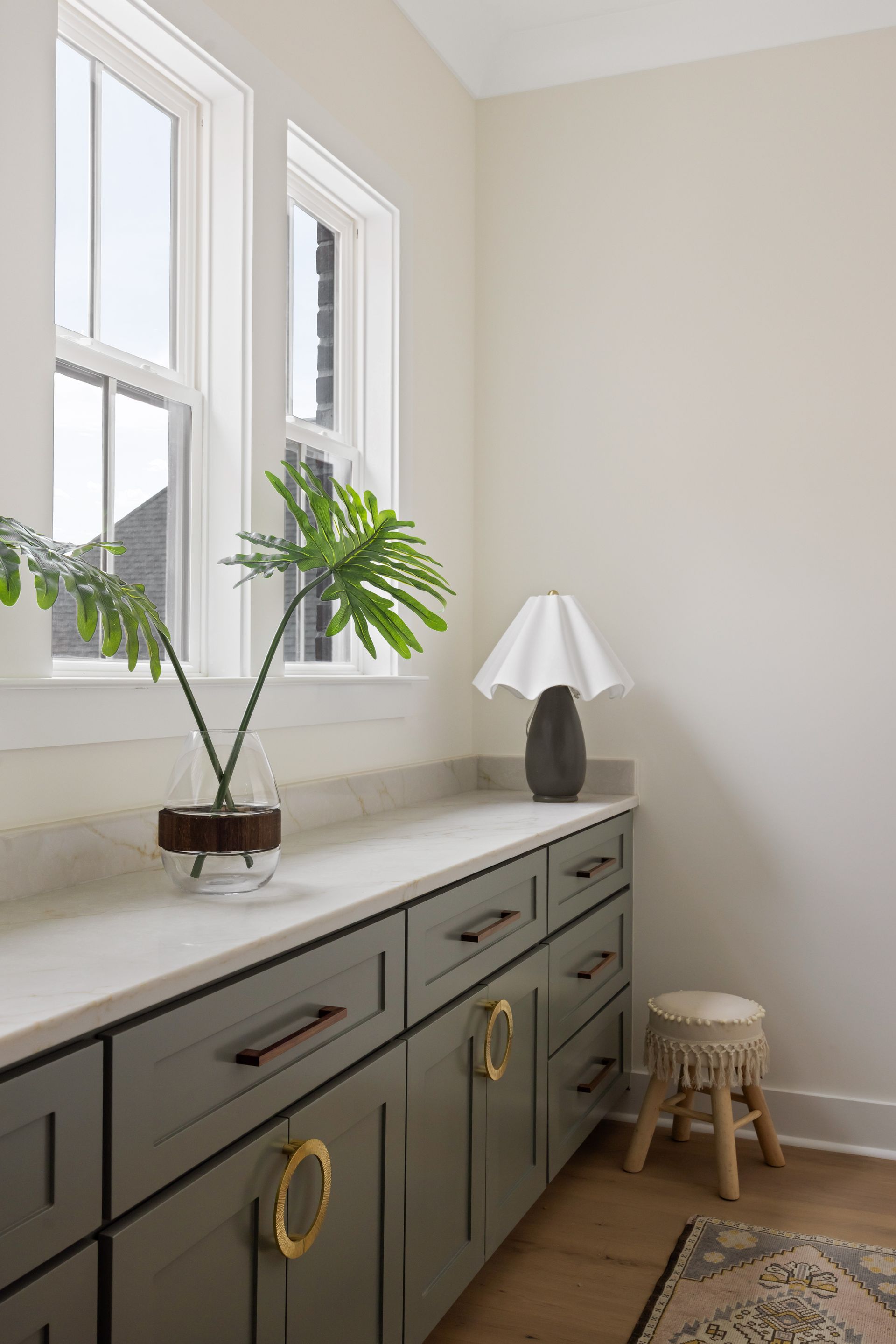 Built-in gray cabinetry with marble countertop, windows, lamp, stool, and plant.