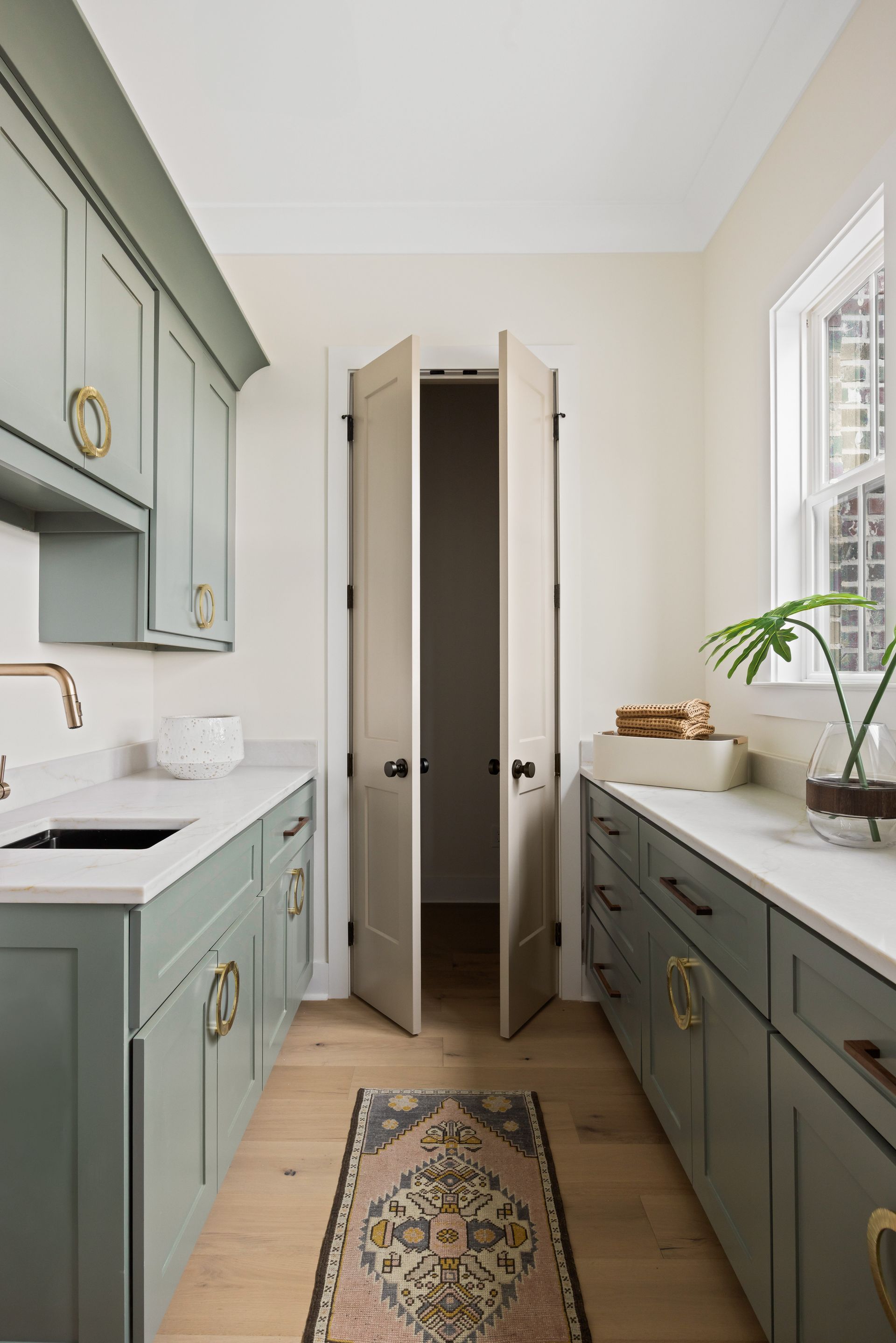 Sage green cabinets line a narrow laundry room. Bi-fold doors are open, rug on the floor. White countertops and light walls.