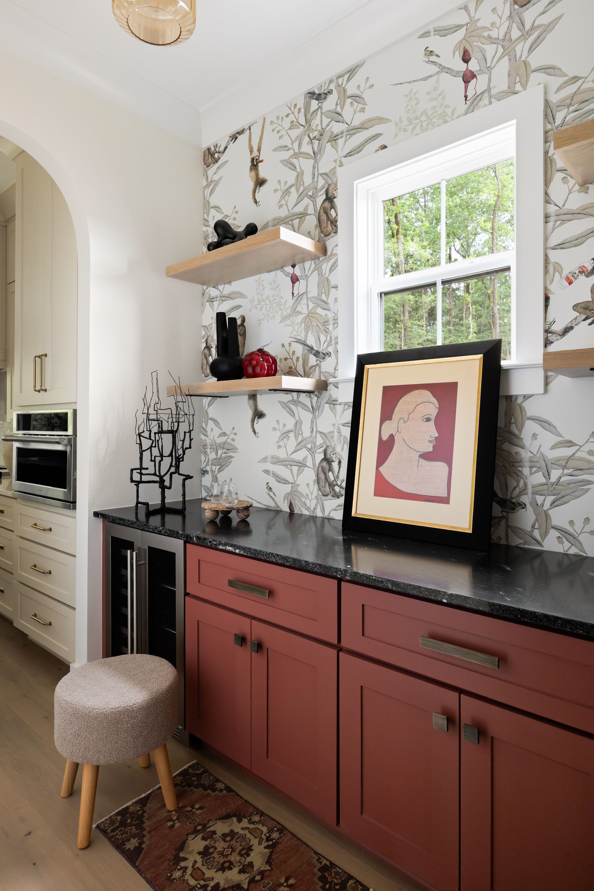 A home bar with red cabinets, a black countertop, and floral wallpaper. There's a small stool and art on display.