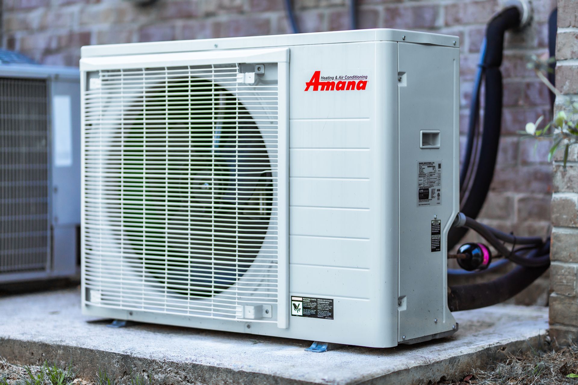 A gray residential air conditioning unit sits on a concrete pad outside a house with light-colored siding and gravel ground.