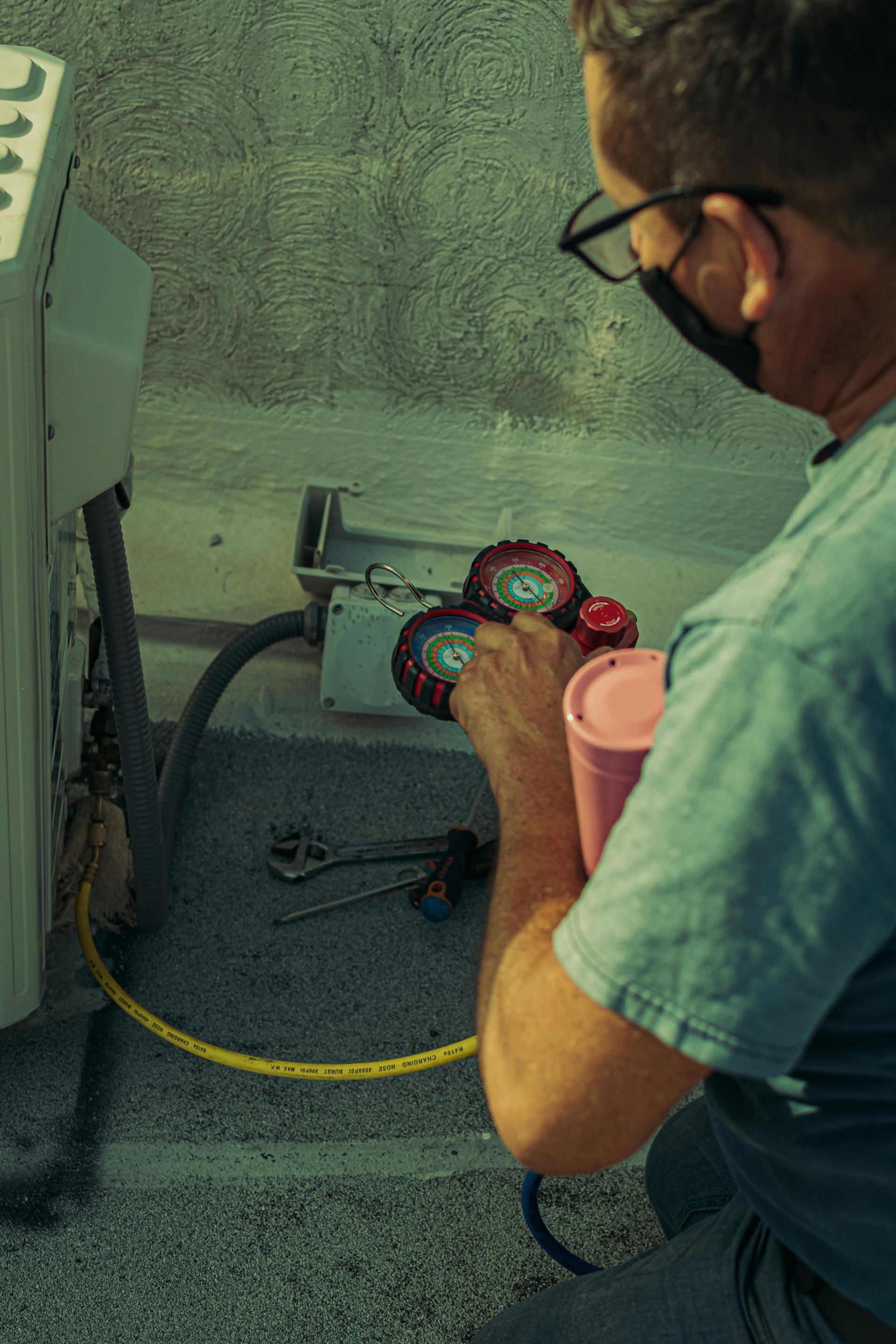 A person in a mask and glasses holds a set of HVAC pressure gauges while working on an outdoor air conditioning unit.