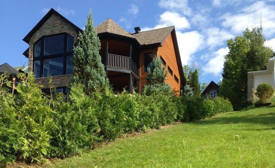 Maison en bois avec façade en pierre, balcon et grandes fenêtres, sur une colline herbeuse sous un ciel bleu.
